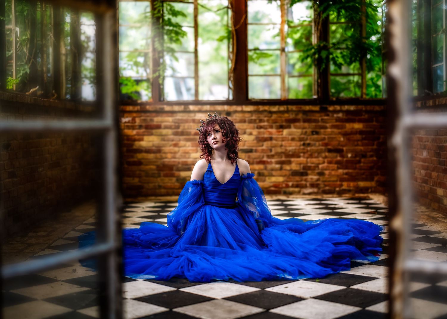 Woman in blue gown seated on checkered floor in greenhouse with brick walls and large windows.