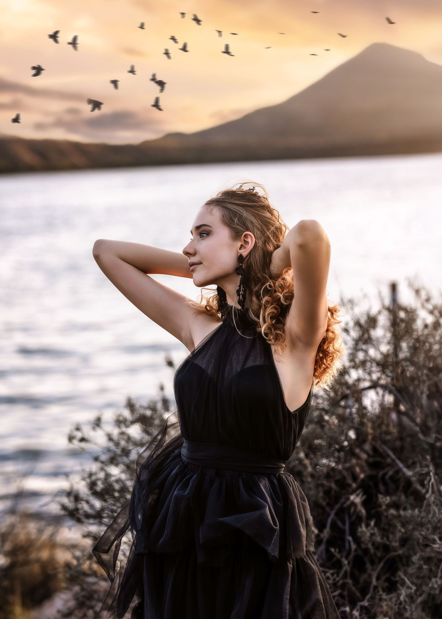 Woman in black dress poses by lake at sunset with birds flying and mountain backdrop.