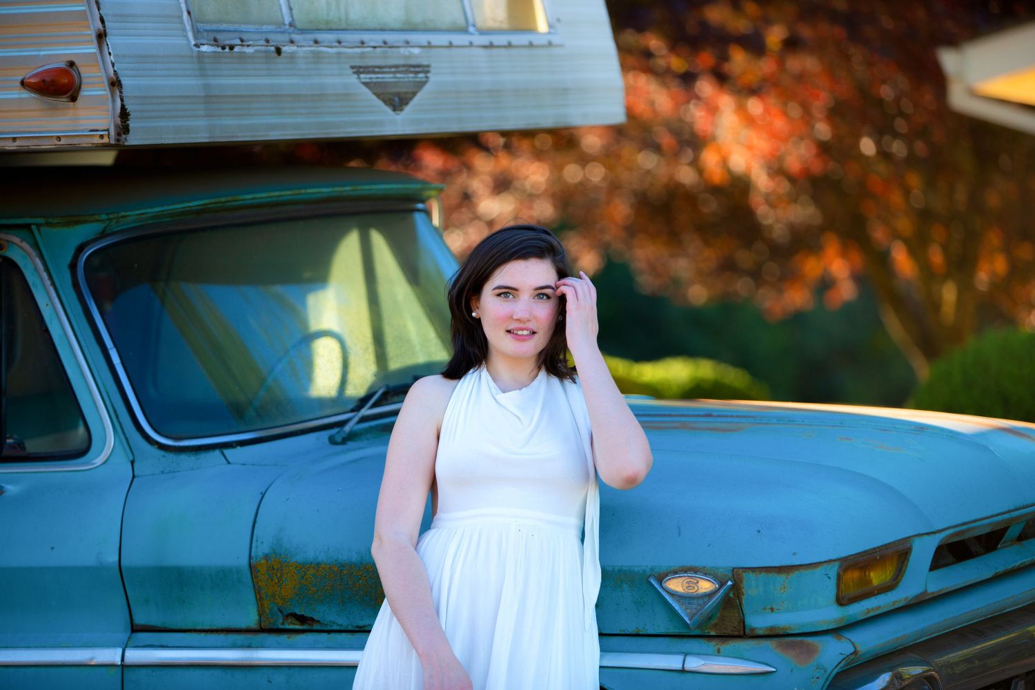 Young woman in white dress adjusting sunglasses by vintage blue truck with autumn foliage.