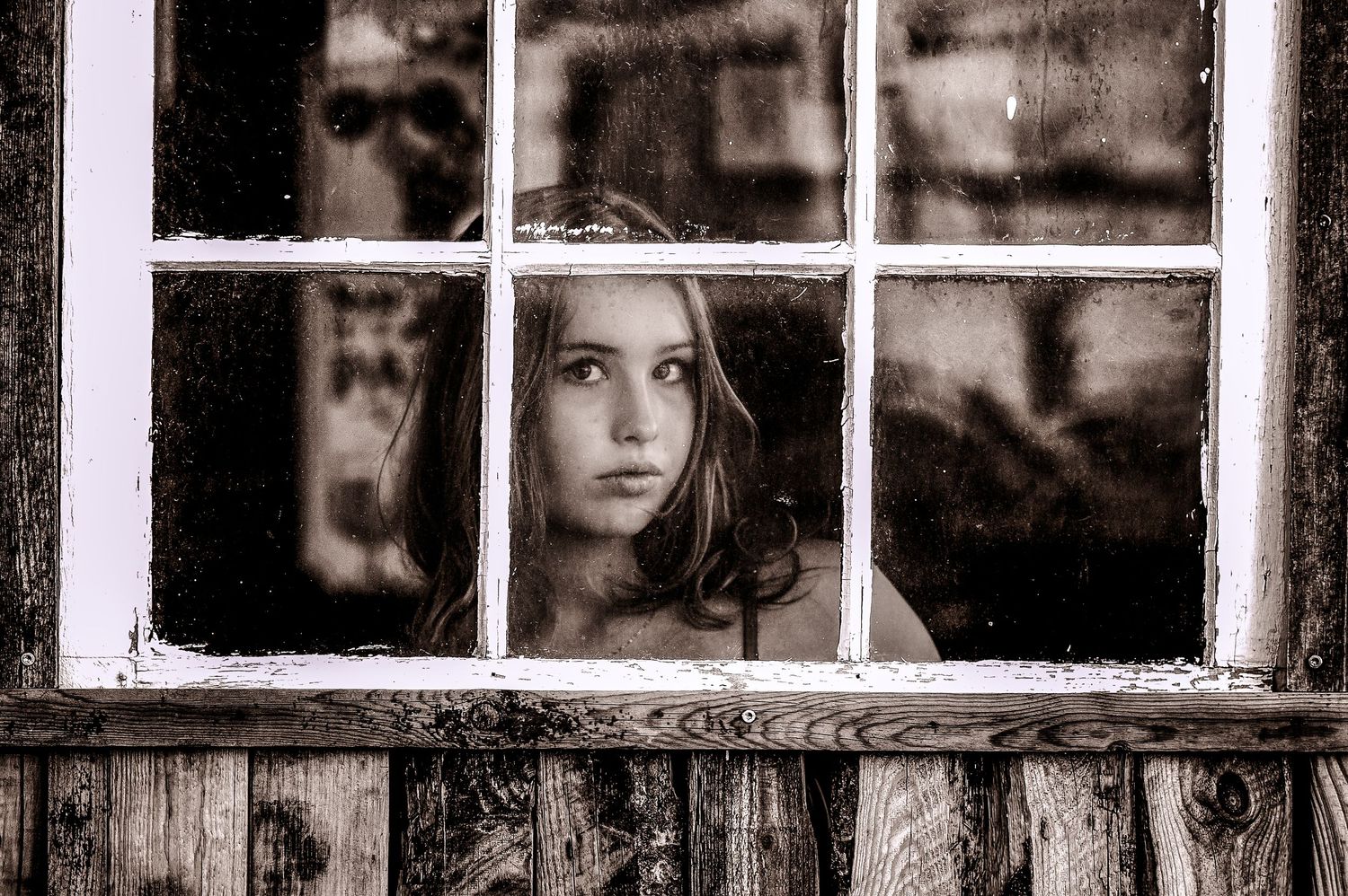 Black and white portrait of young girl gazing through vintage wooden window panes pensively.