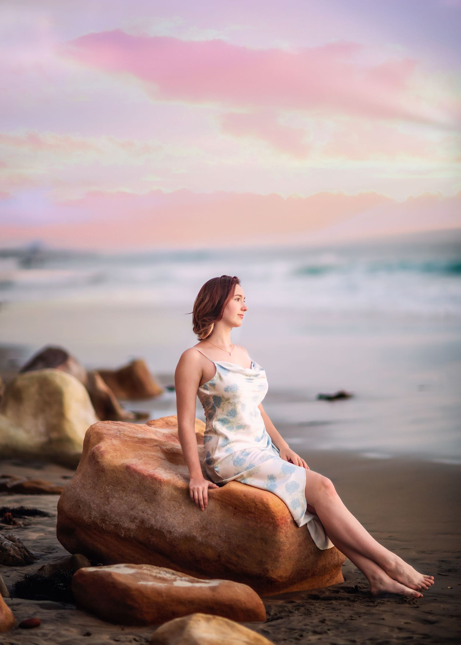 Woman in floral dress sitting on beach rocks at sunset.