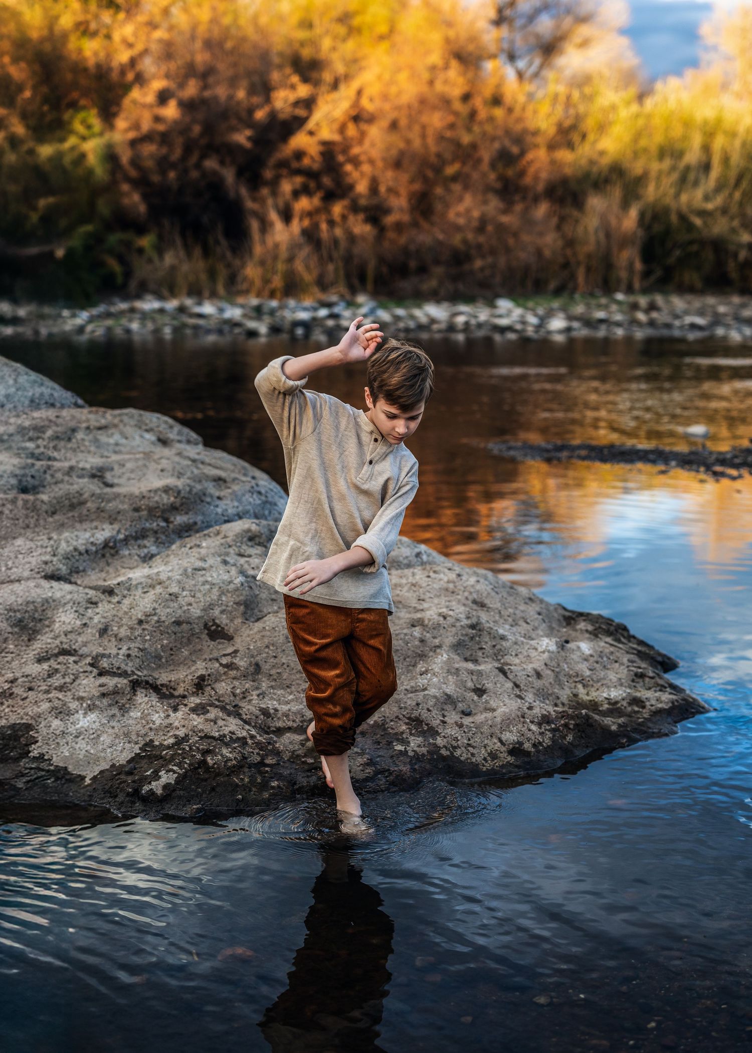Boy wading barefoot in shallow river near large rock with golden autumn trees reflecting in water.