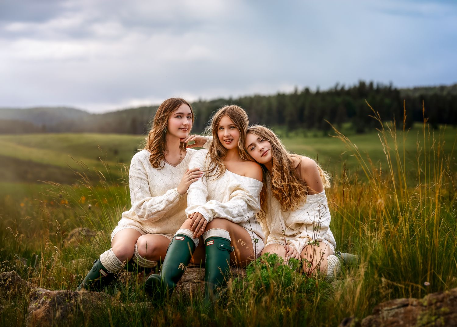 Three young women in cream sweaters and green boots sit together in a lush green field with misty hills behind them.