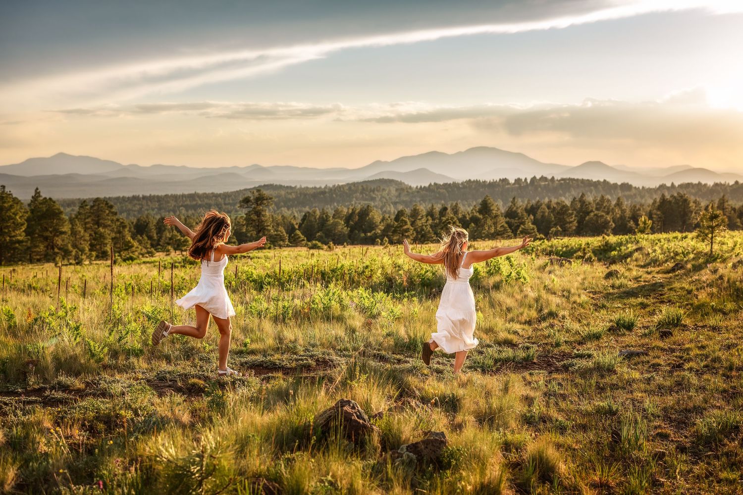 Two girls in white dresses dance in a sunlit meadow with mountain views and forest backdrop at golden hour.