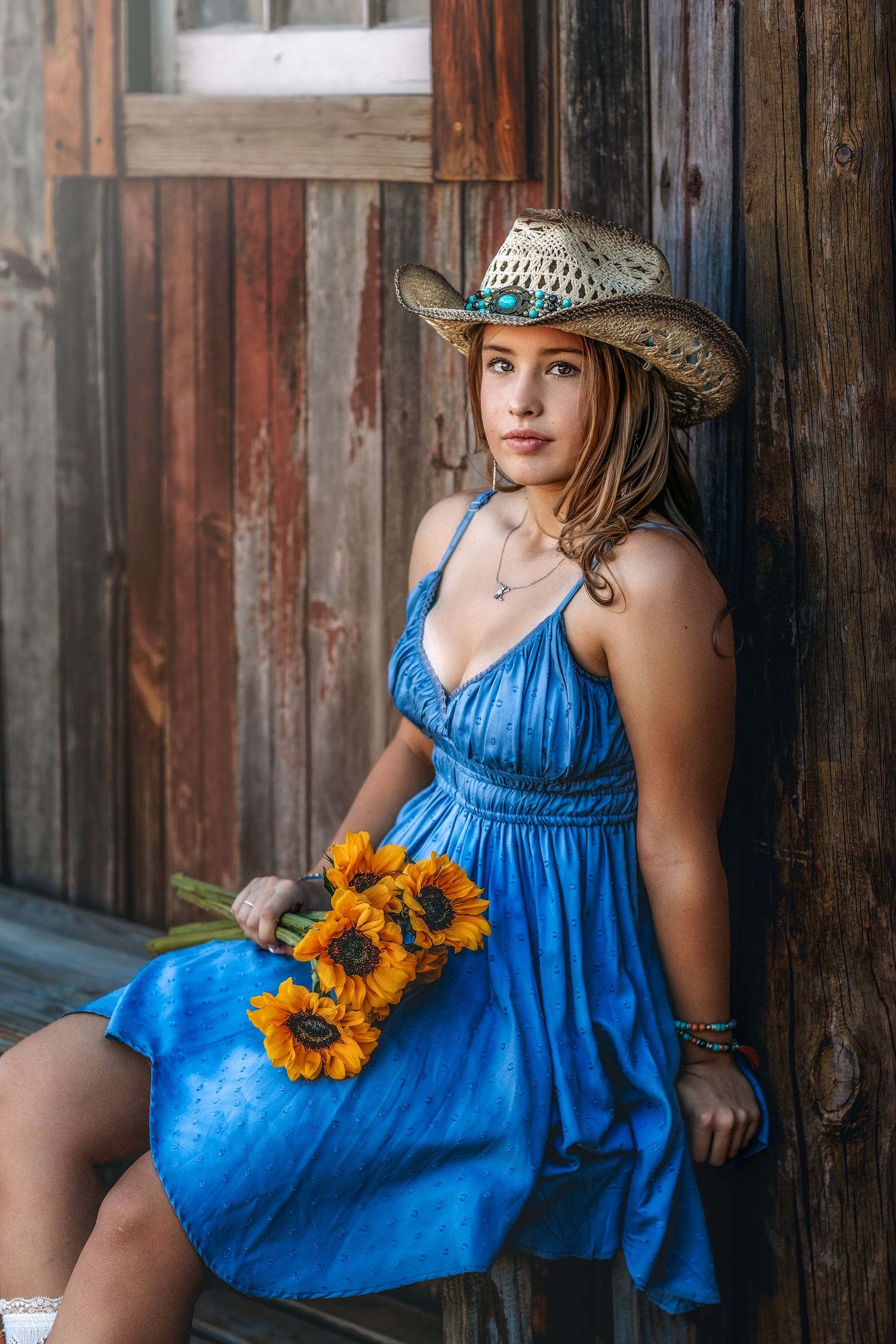 Woman in blue dress and cowboy hat sitting with sunflowers against weathered wood wall.