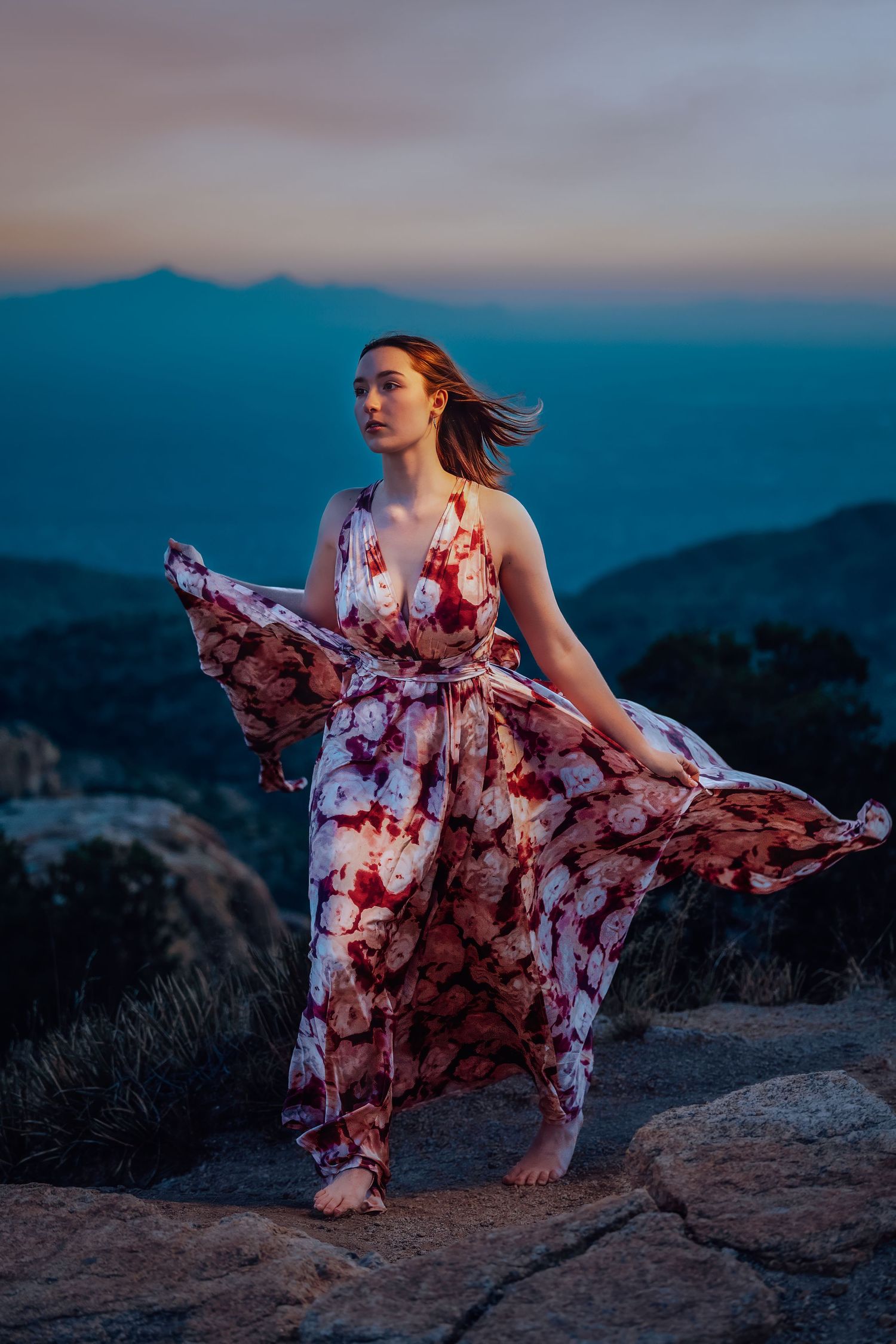 Barefoot woman in flowing floral dress stands on mountain rocks at blue dusk twilight.