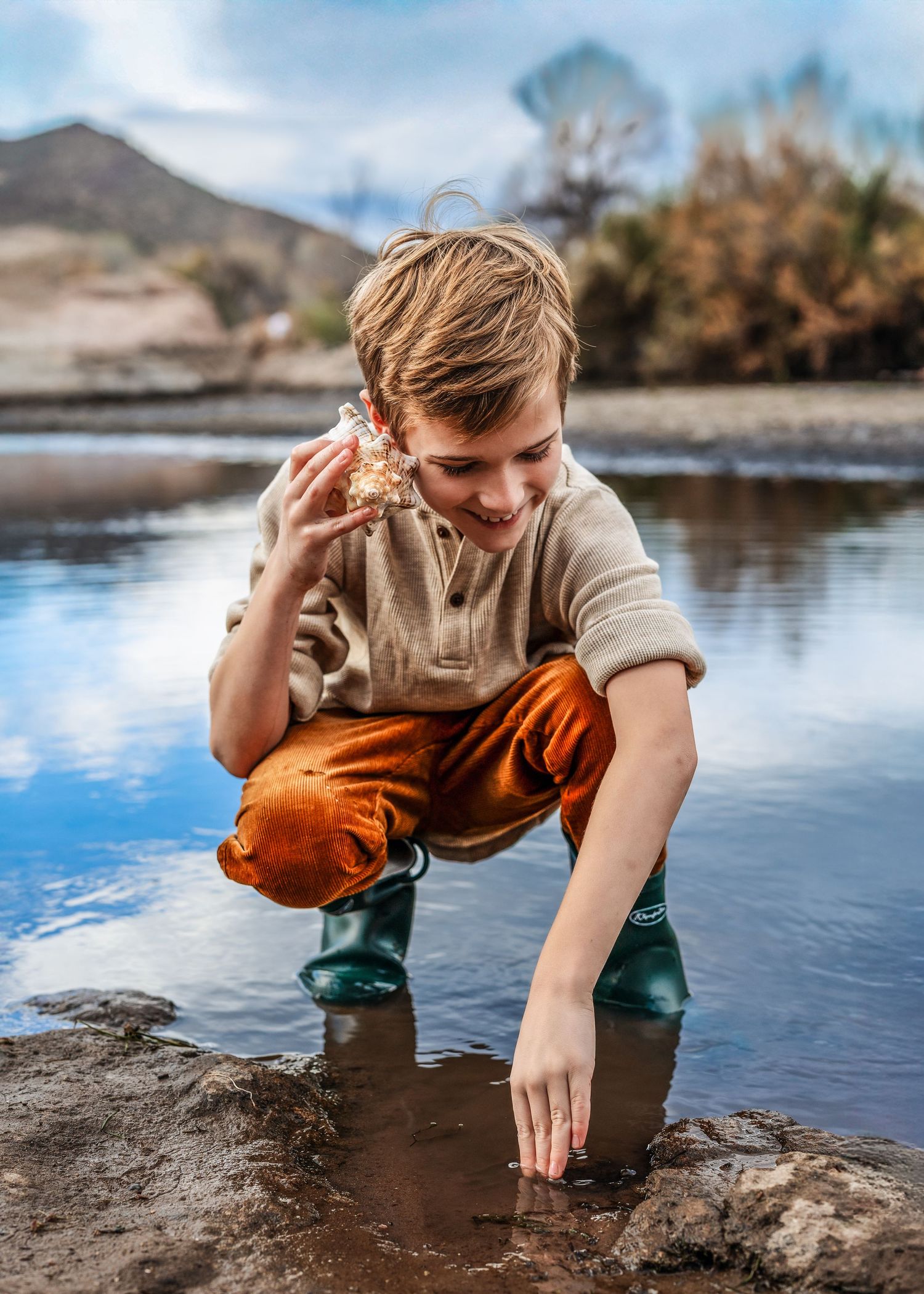Young boy in rubber boots crouches by water examining a rock while exploring a scenic river.