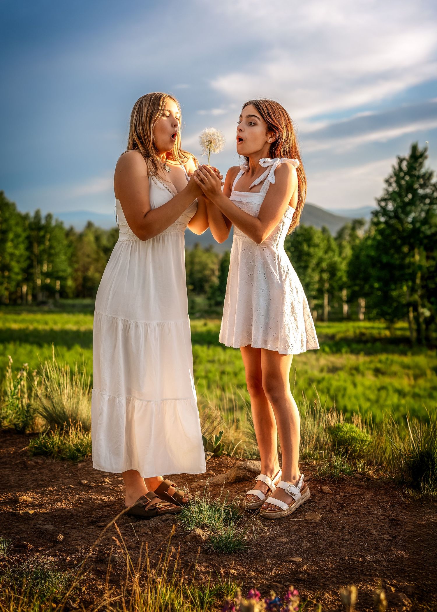 Two young women in white dresses blowing a dandelion together in a sunny green field.