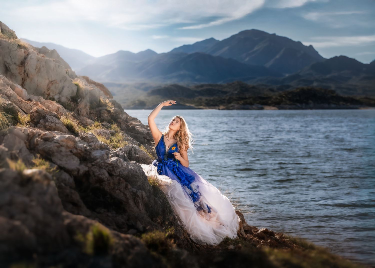Woman in blue and white gown poses gracefully on rocky lakeside with mountain backdrop.