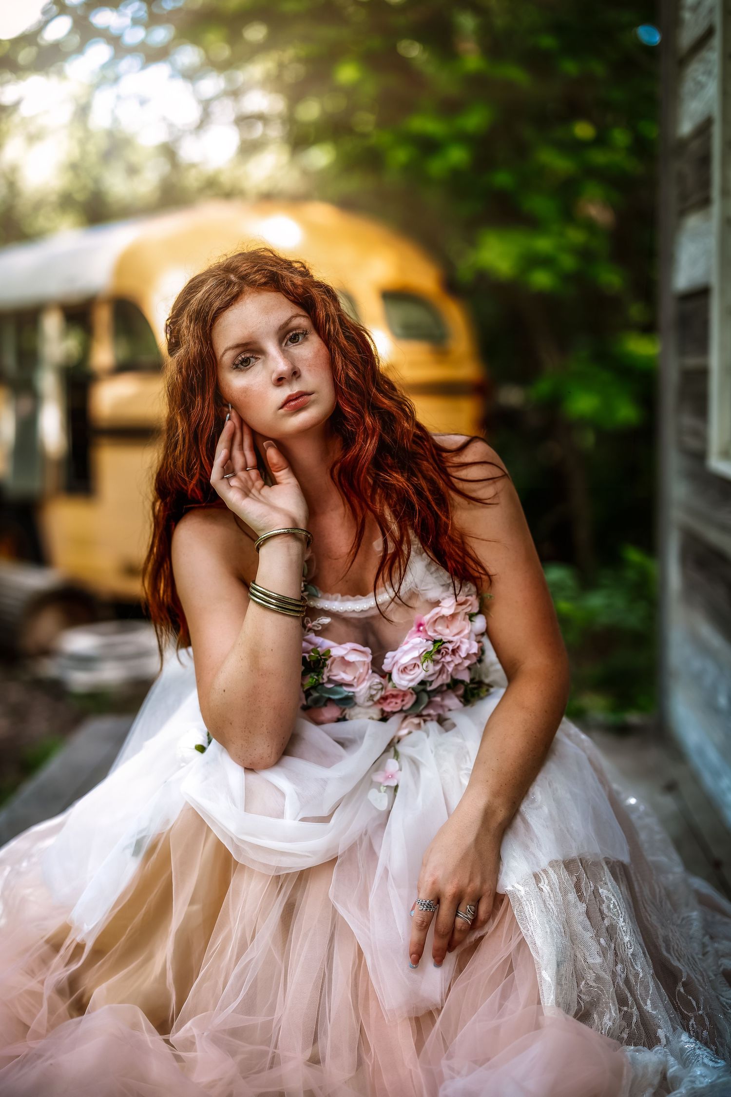Red-haired woman in floral tulle gown poses thoughtfully outdoors near a vintage yellow school bus.