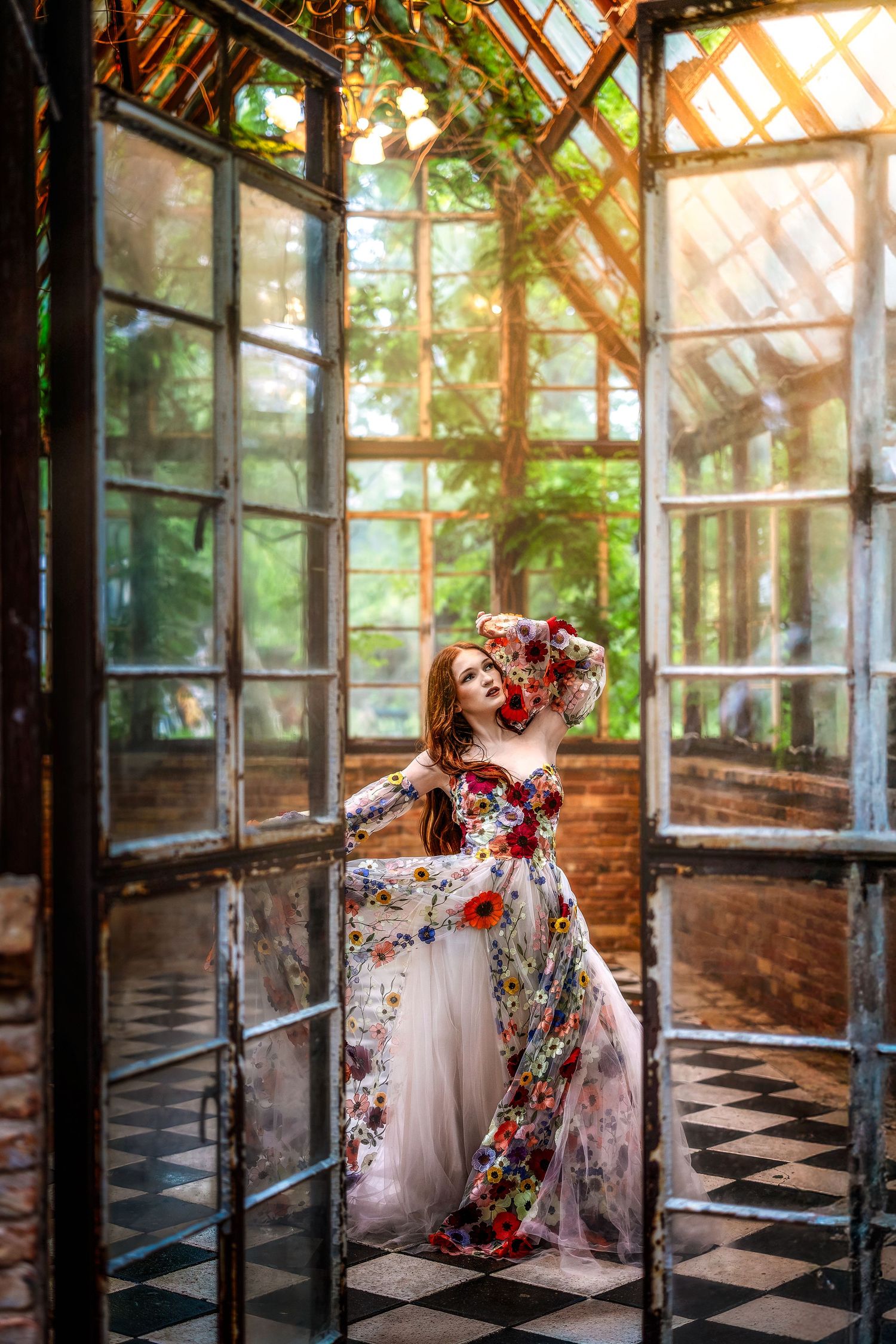 Woman in a floral embroidered white gown poses gracefully inside a sunlit vintage greenhouse with checkered floors.