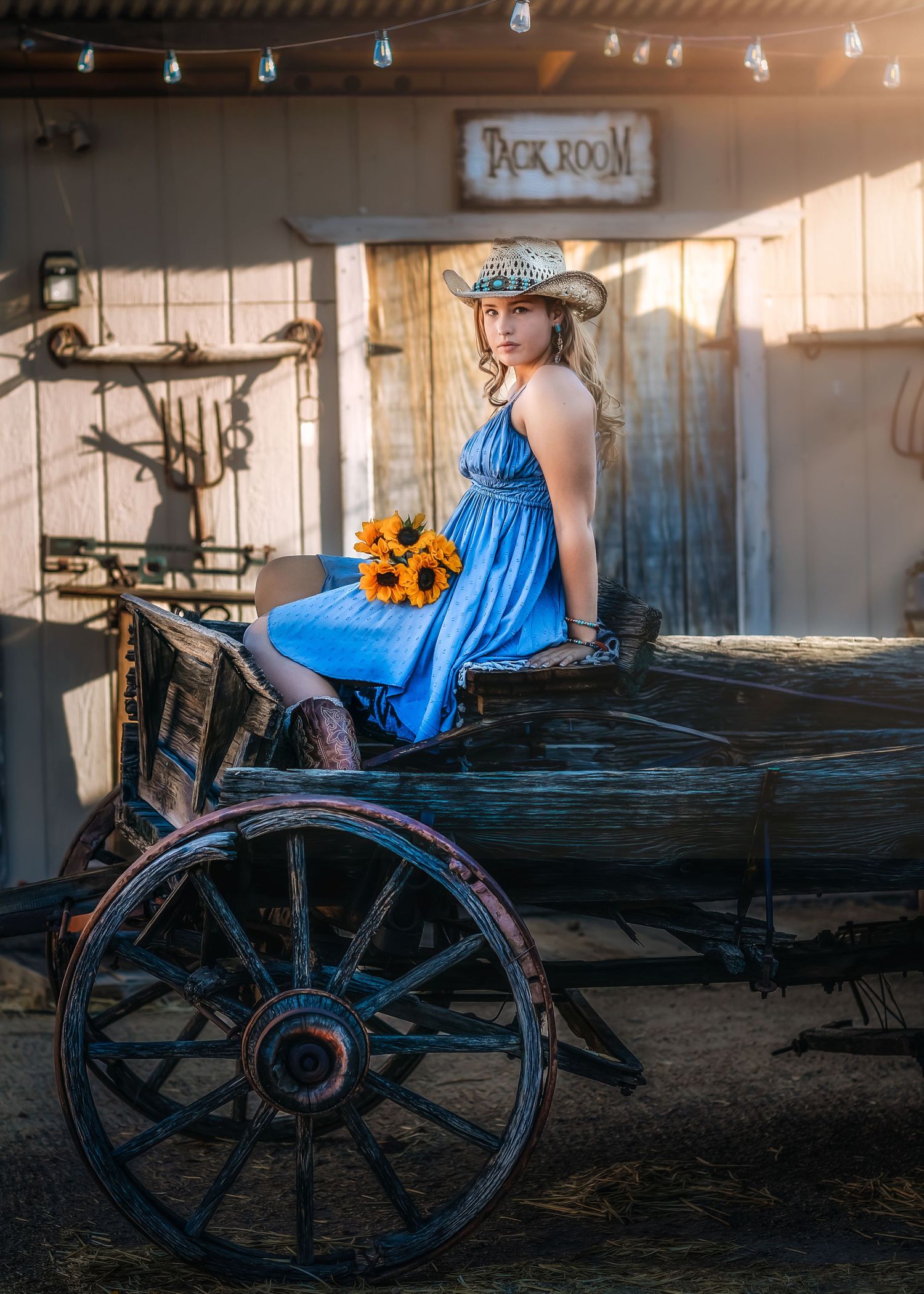 Woman in blue dress and cowboy hat sitting on antique wooden wagon with sunflowers.