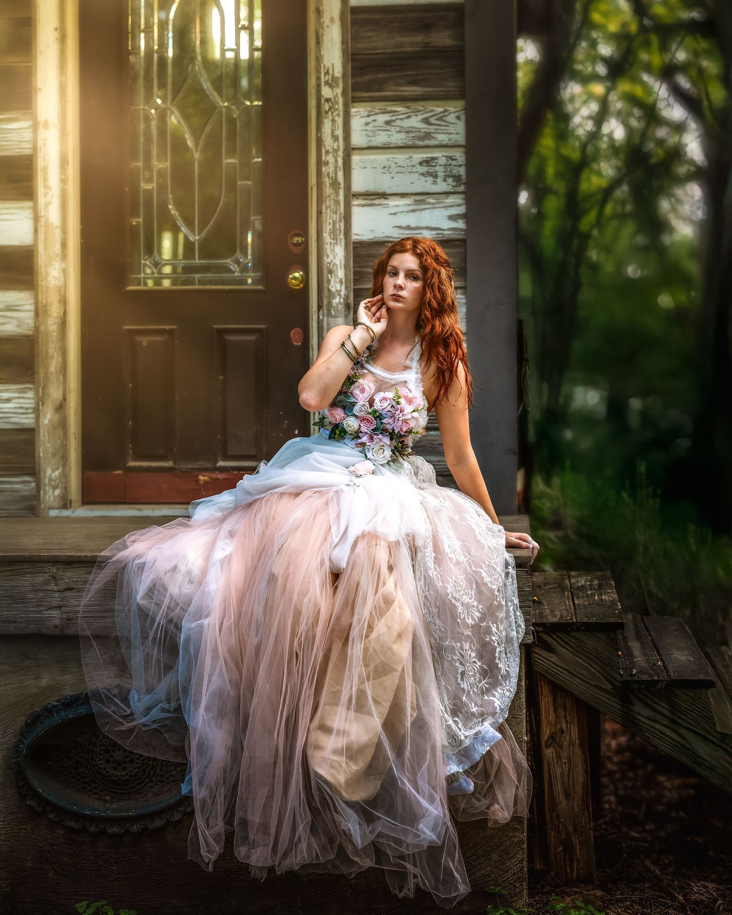 A redhead bride in a flowing pink tulle gown sits on a rustic porch holding a bouquet, bathed in warm golden light.