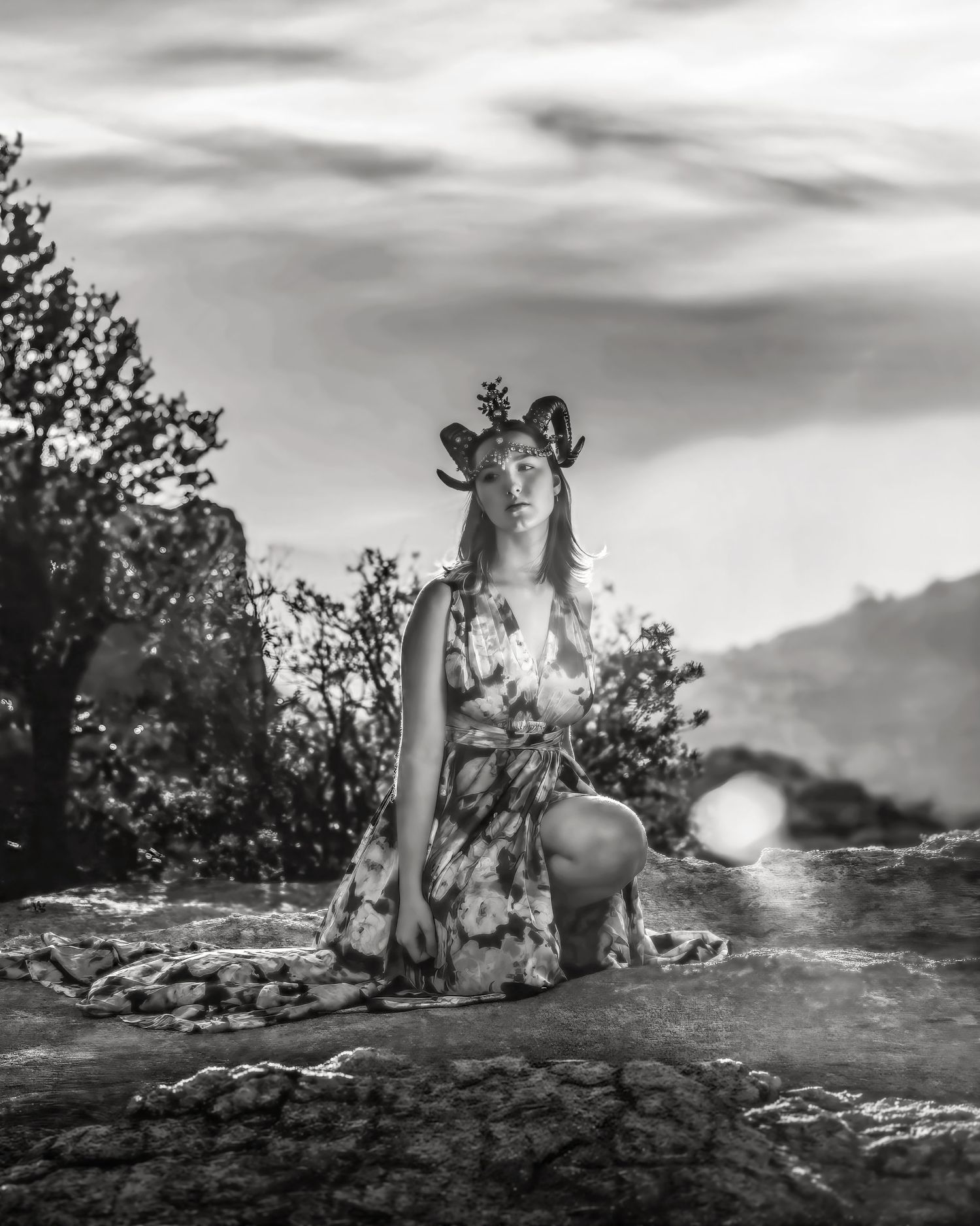 Black and white photo of woman in floral dress with antler crown kneeling on rocky terrain.