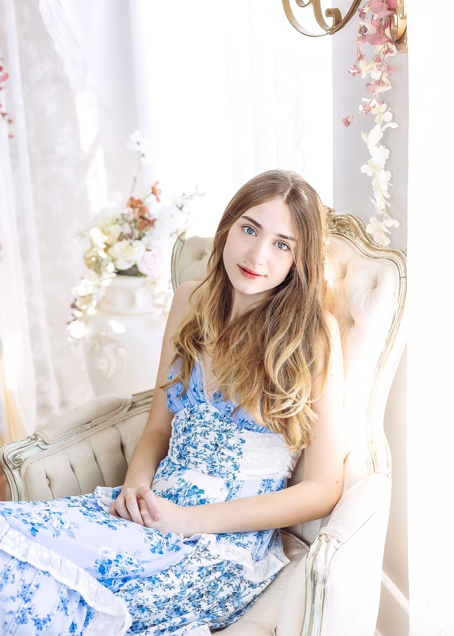 Young woman with long hair in blue floral dress seated in cream French armchair by window.