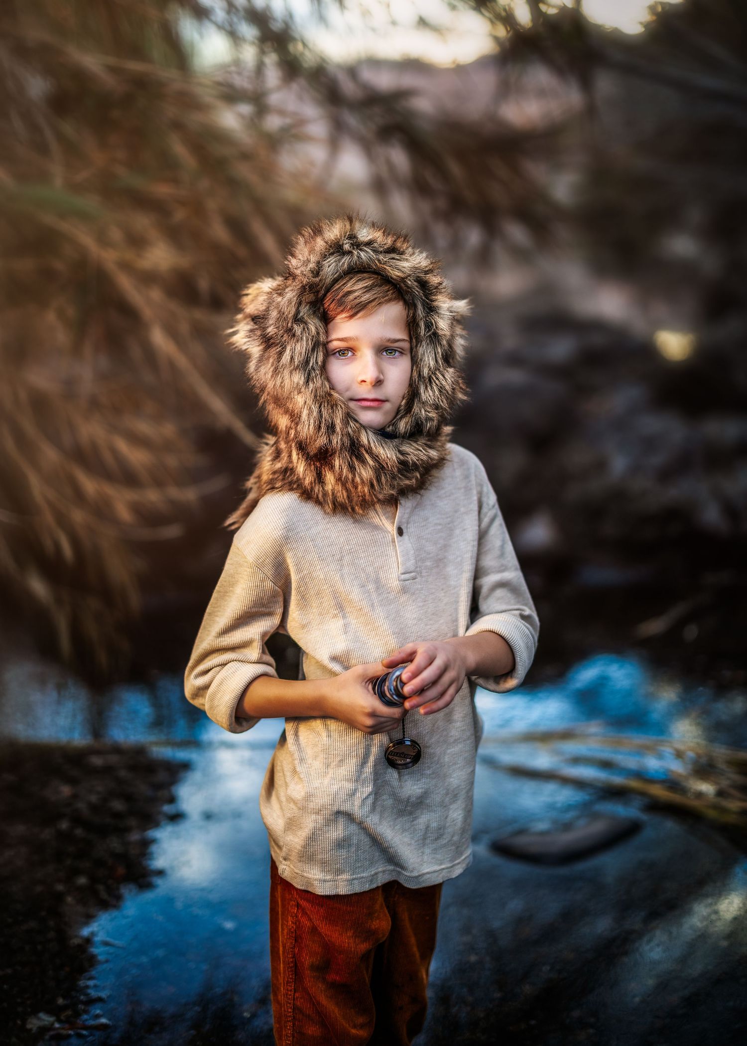 Child wearing fur-trimmed hood holding small object standing near icy stream in nature.