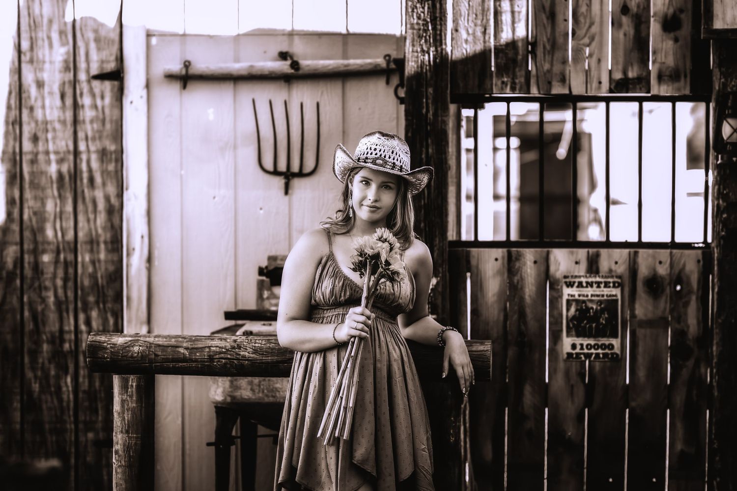 Sepia-toned portrait of woman in cowboy hat holding flowers in vintage western barn setting.