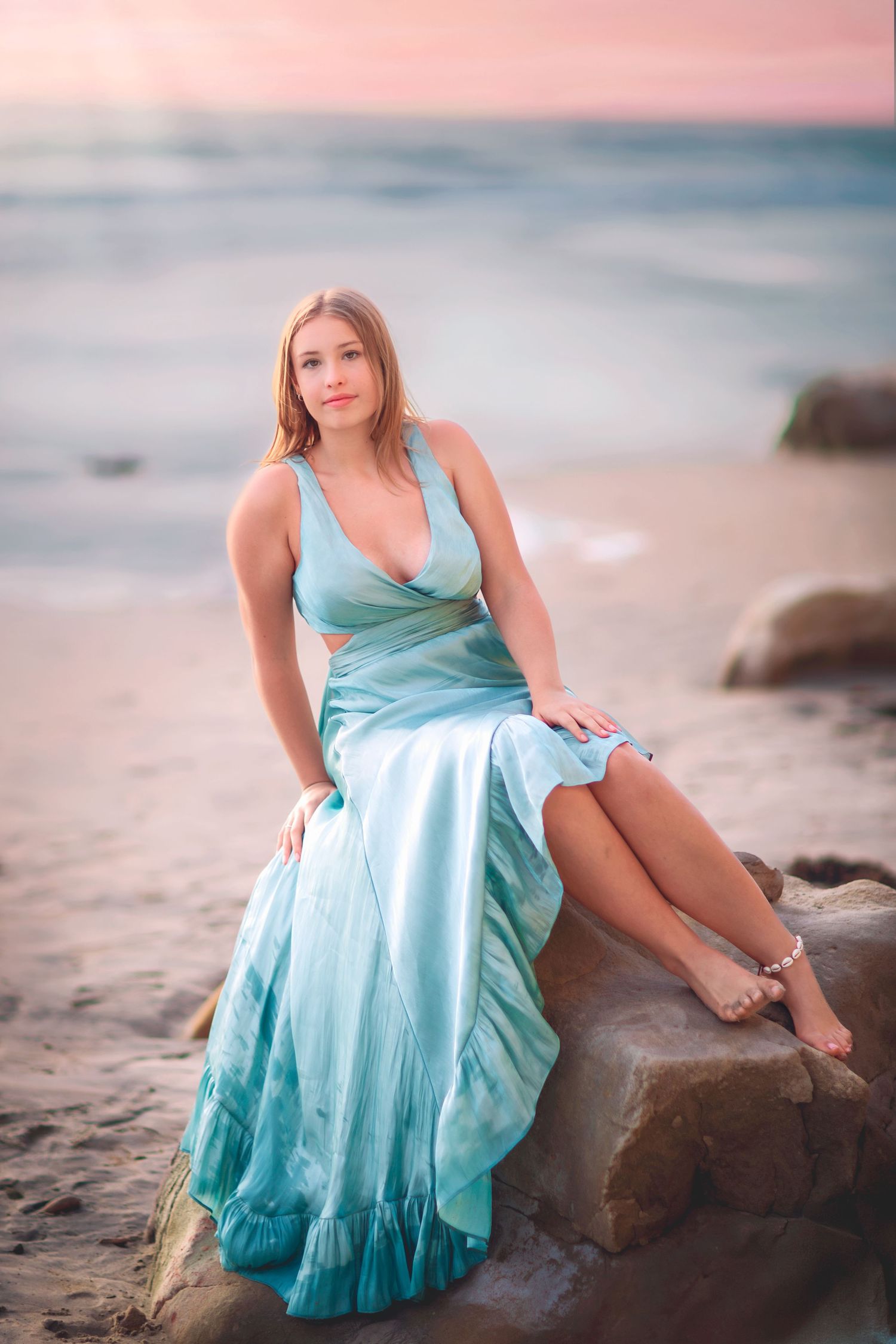 Blonde woman in teal evening gown sits on beach rocks during colorful pastel sunset.