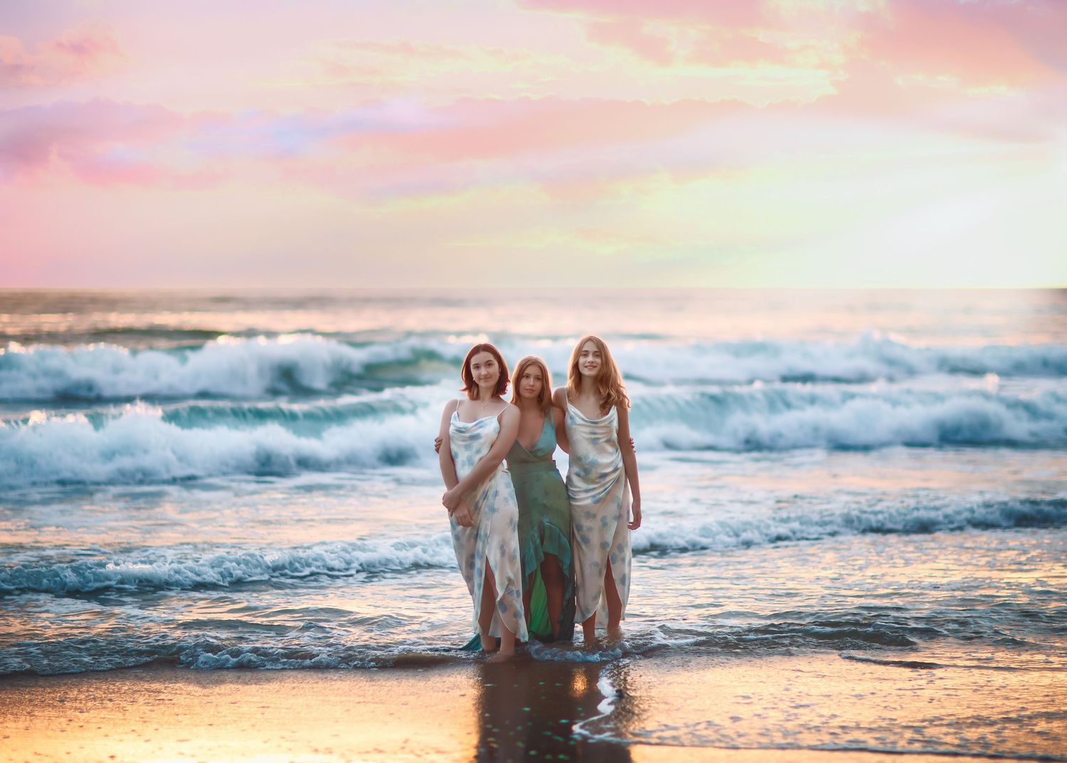 Three women in elegant dresses standing on beach at sunset.
