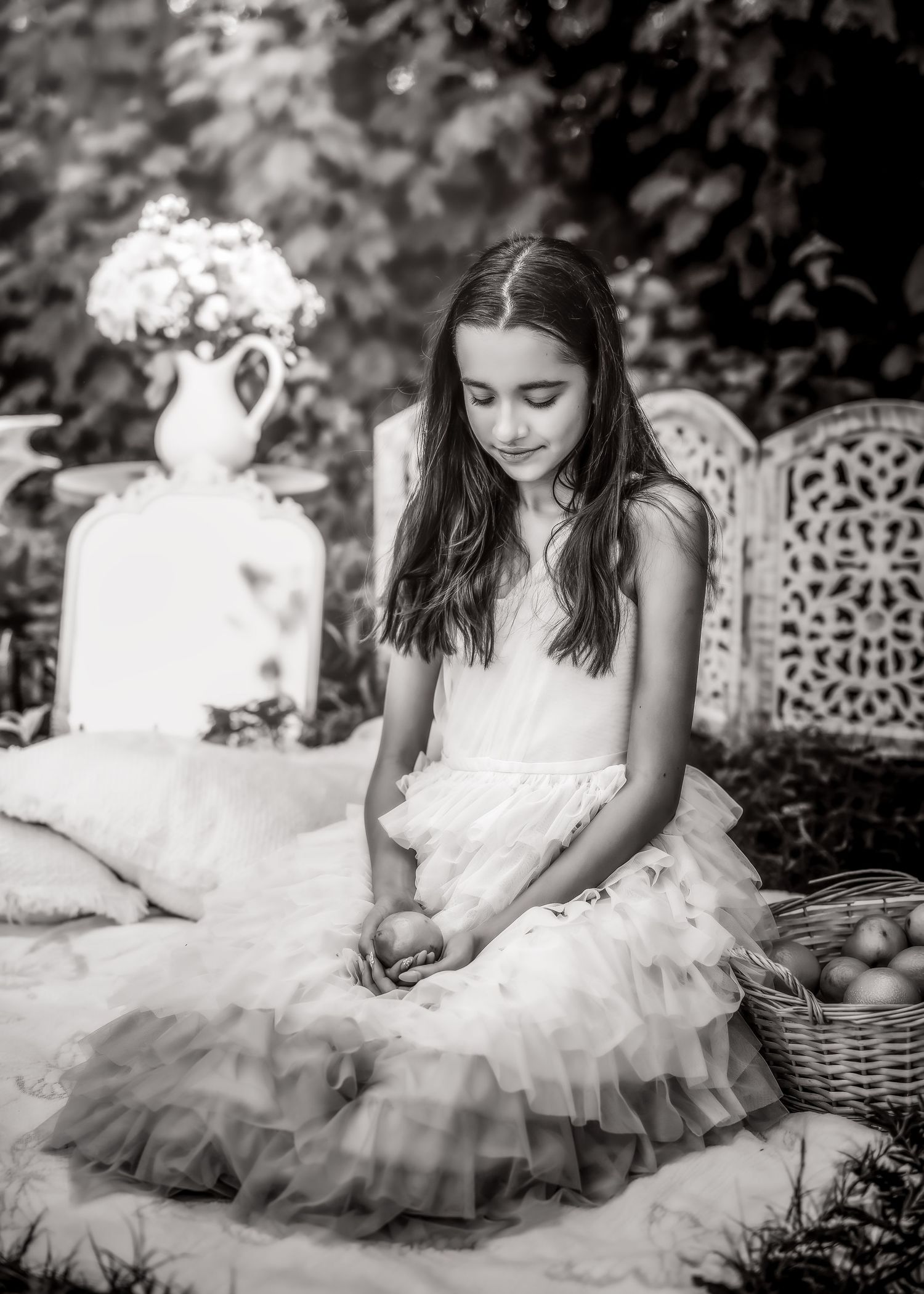 Black and white photo of a girl in a white tulle dress sitting on a blanket holding fruit in a garden.