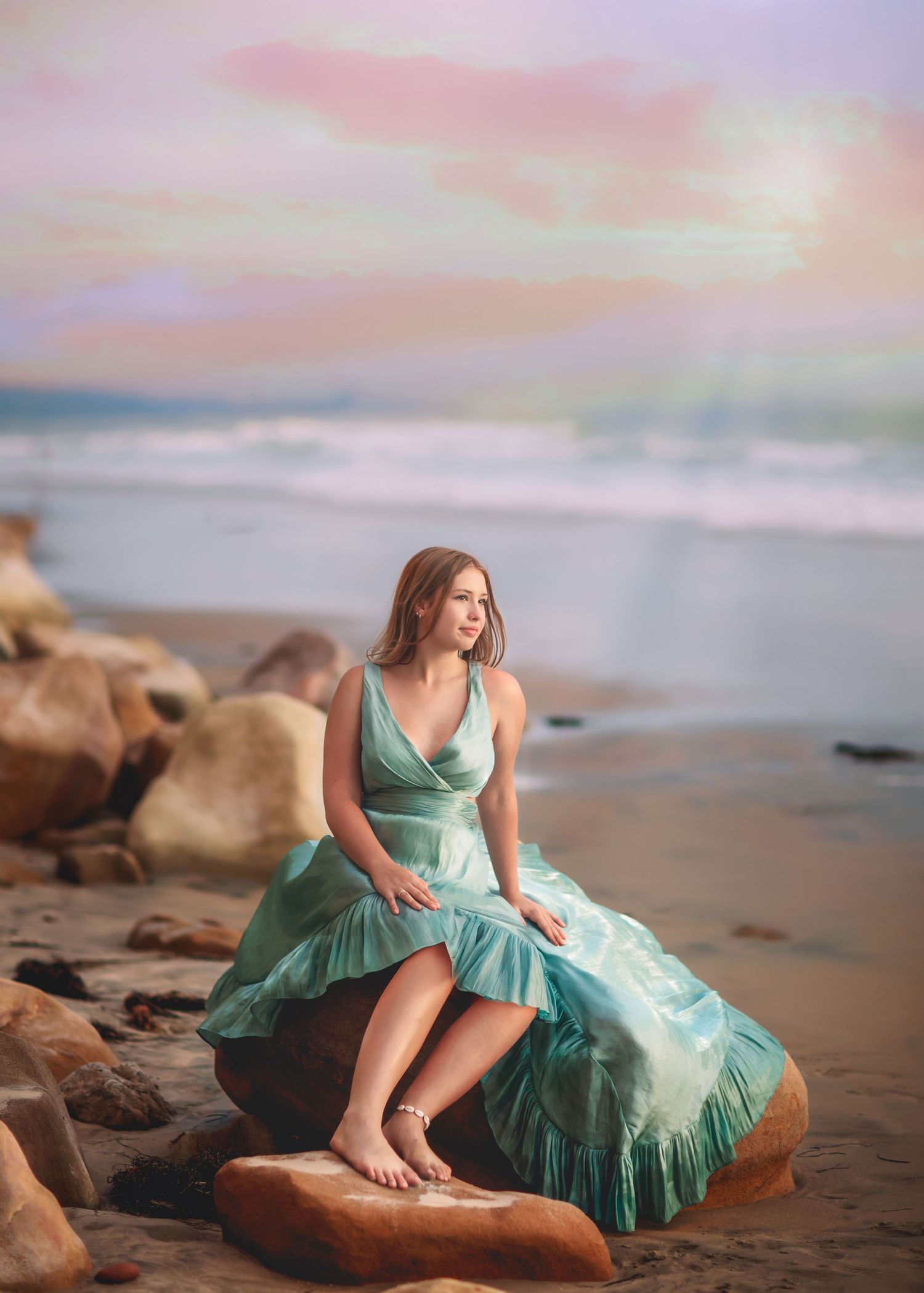 Young woman in teal gown sits gracefully on beach rock against dreamy pastel sunset sky.