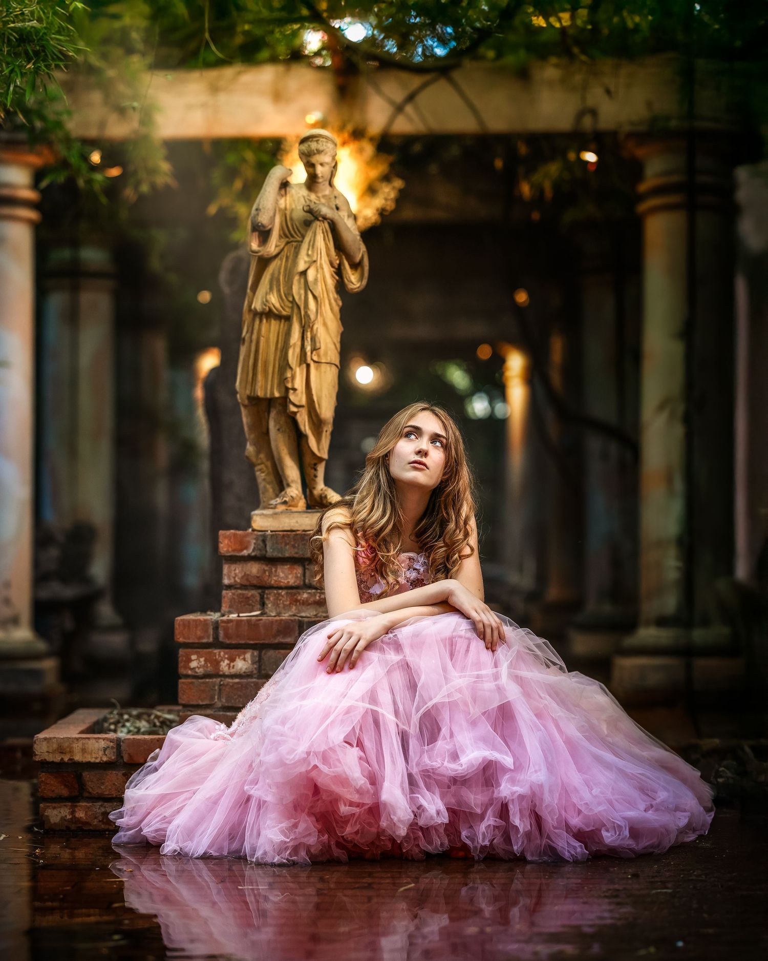 Young woman in pink tulle gown sits gracefully before a classical statue in an enchanting garden setting.