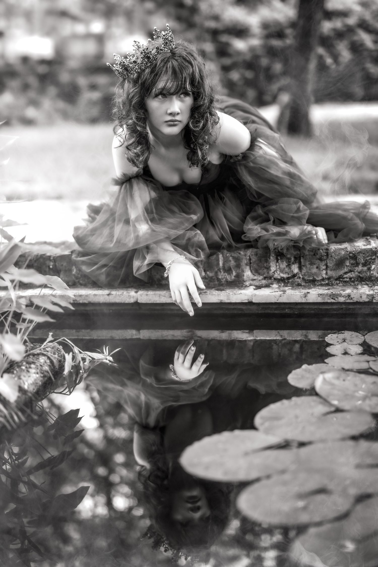Black and white photo of a woman in a flowing dress with flower crown, gazing at her reflection in a lily pond.