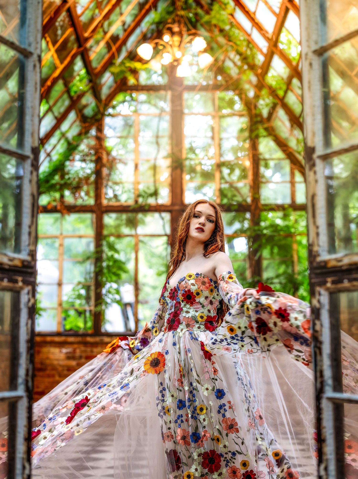 Woman in floral gown twirling in a sunlit greenhouse with lush greenery and arched glass windows.
