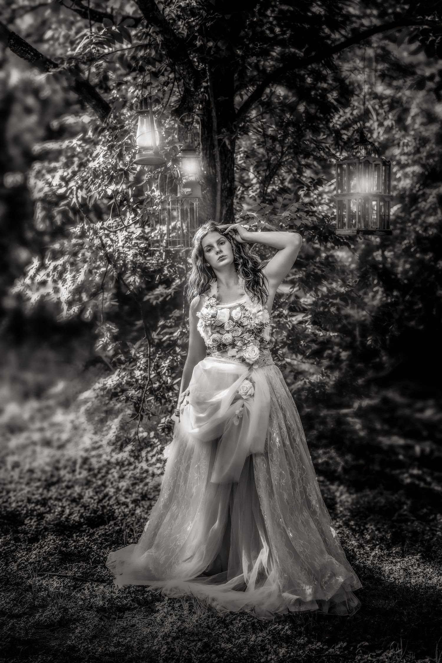 Black and white photo of a bride in a flowing gown holding flowers, standing among trees with glowing lanterns.