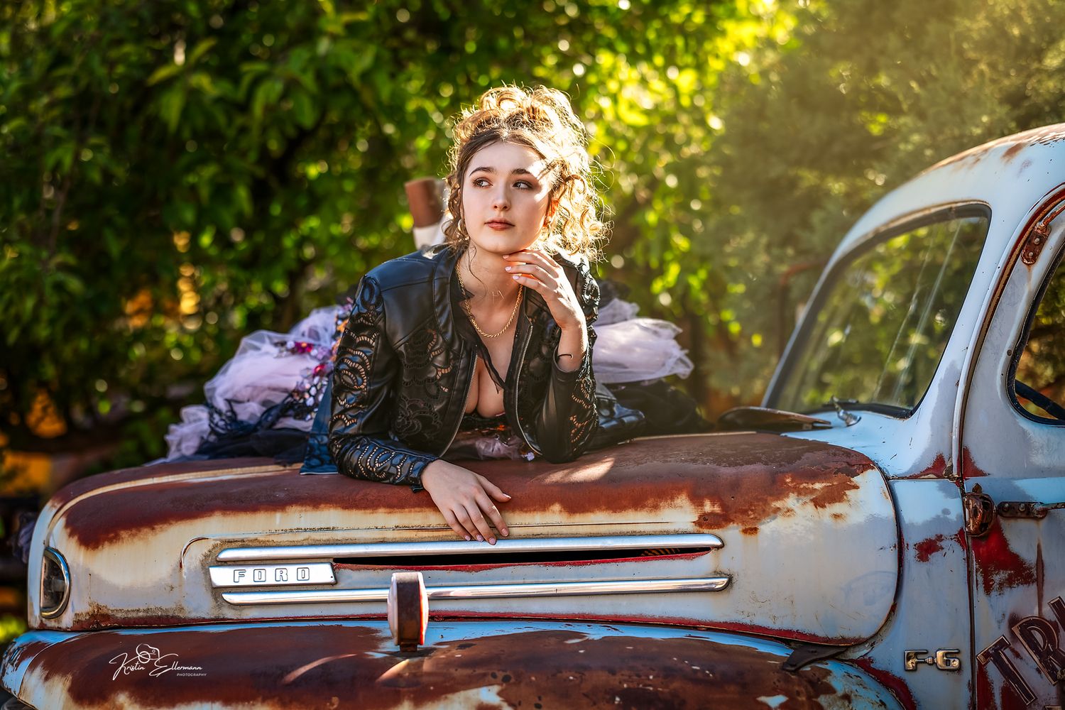 Woman in black lace jacket poses on vintage rusty car hood outdoors with lush green trees in background.
