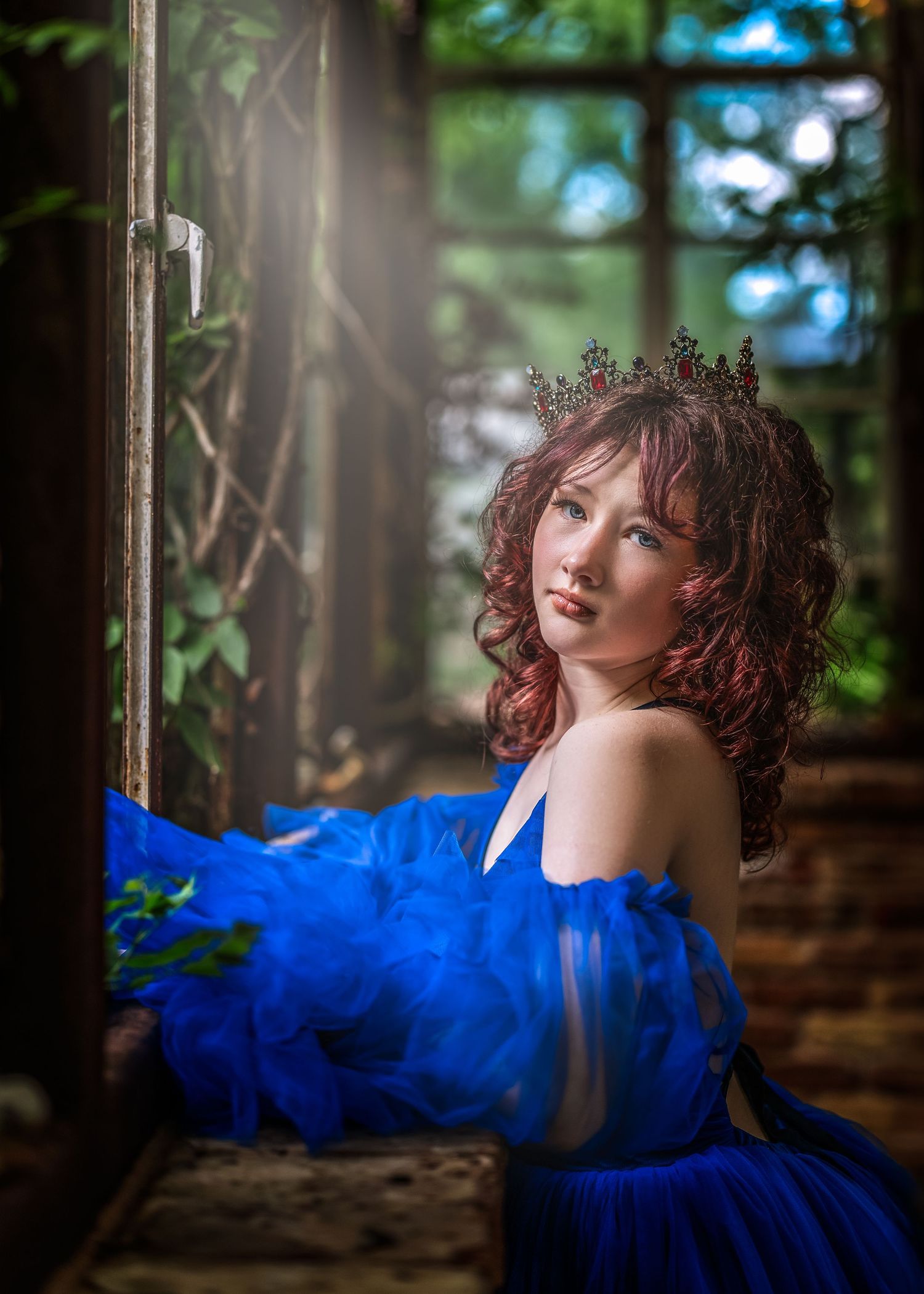 Young woman in a royal blue tulle gown and crown gazes upward near a rustic window surrounded by lush greenery.