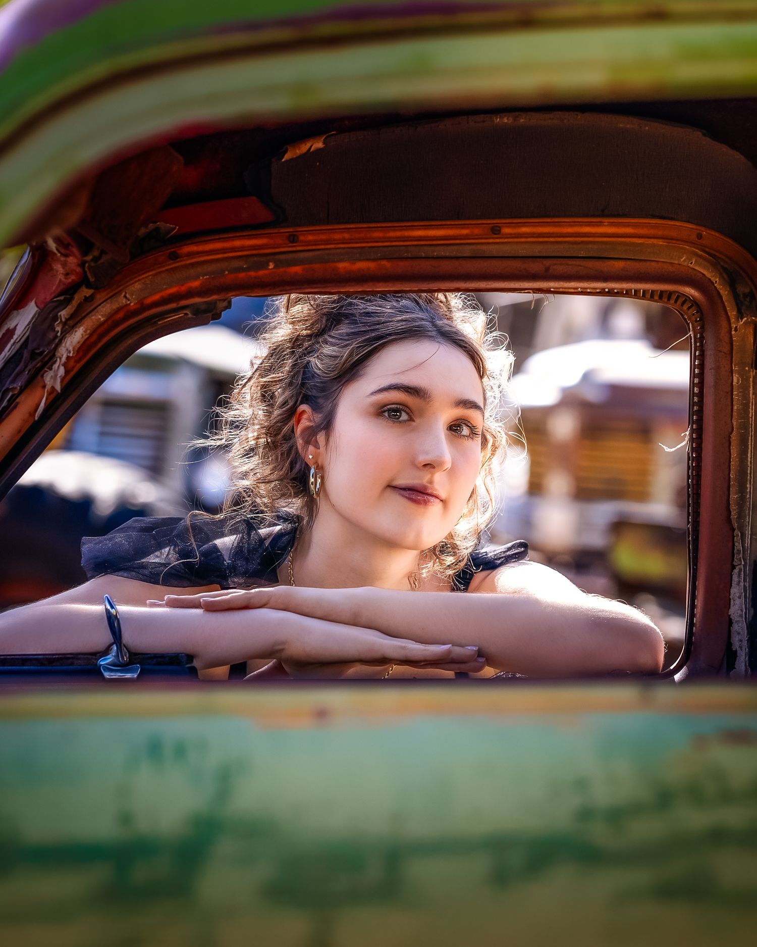 Young woman resting arms on rustic vintage car window, looking contemplatively at camera outdoors.