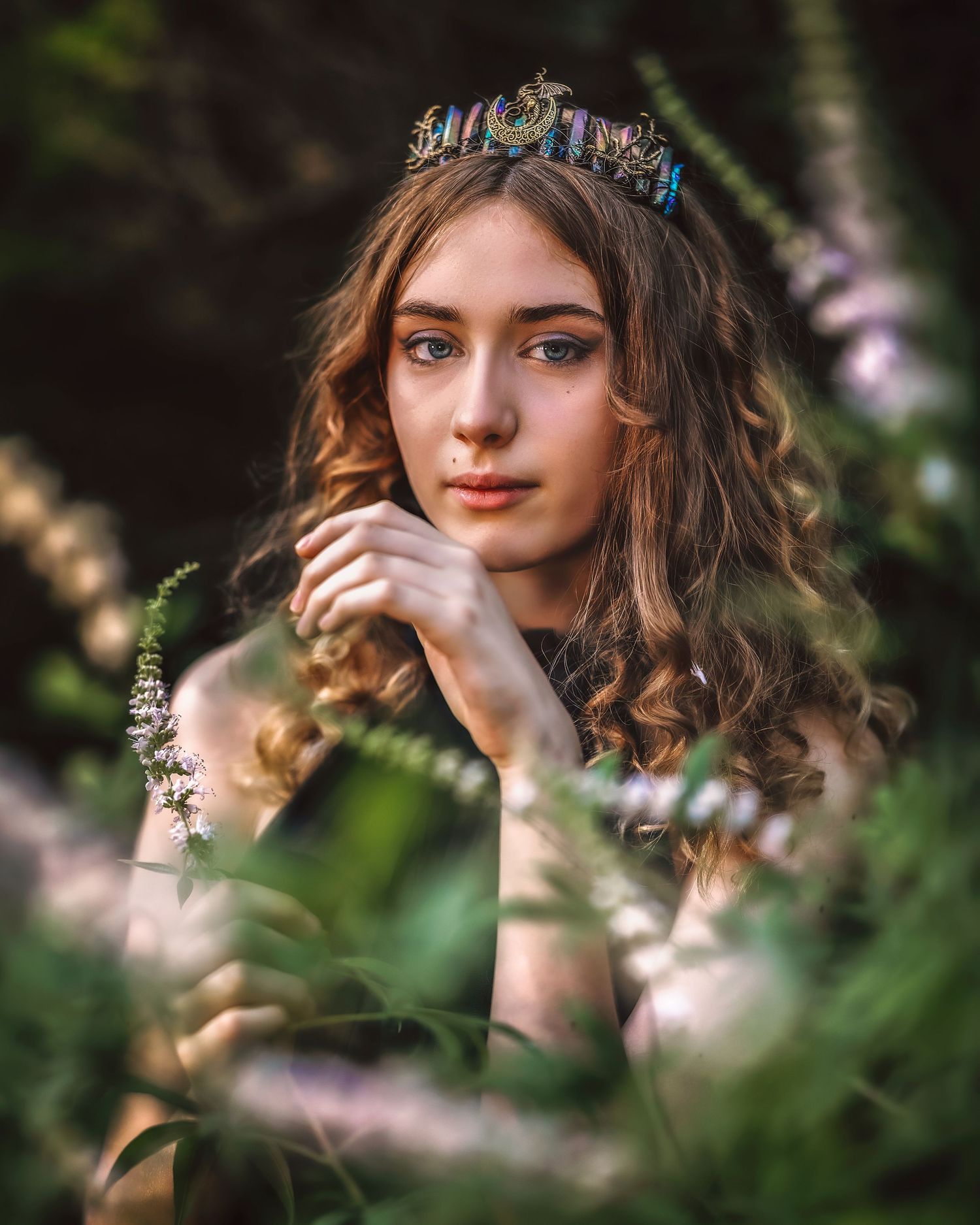 Young woman wearing a decorative crown poses among lush green plants and purple flowers in a dreamy outdoor portrait.