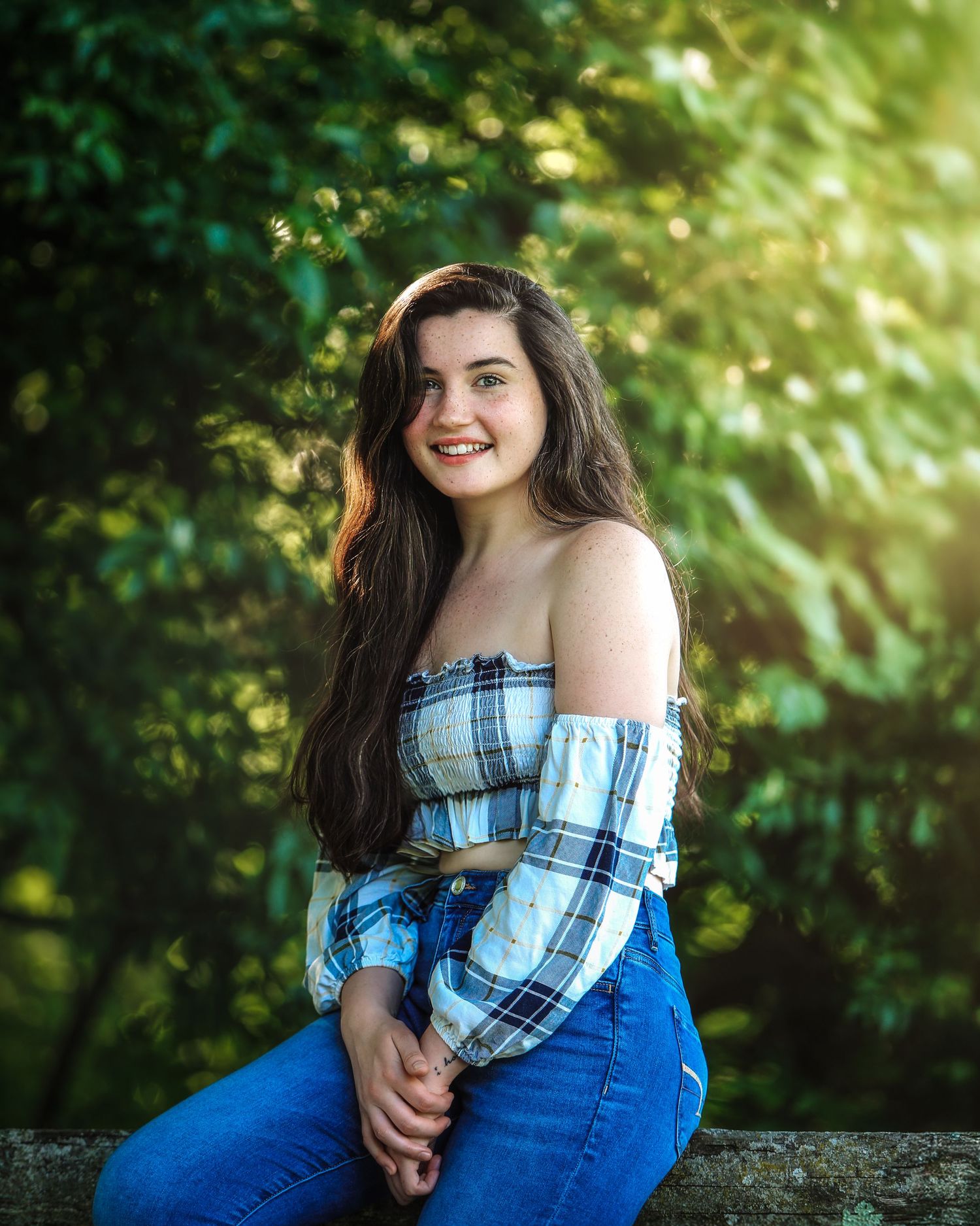 Smiling teenage girl in plaid off-shoulder top and jeans sitting against green foliage.