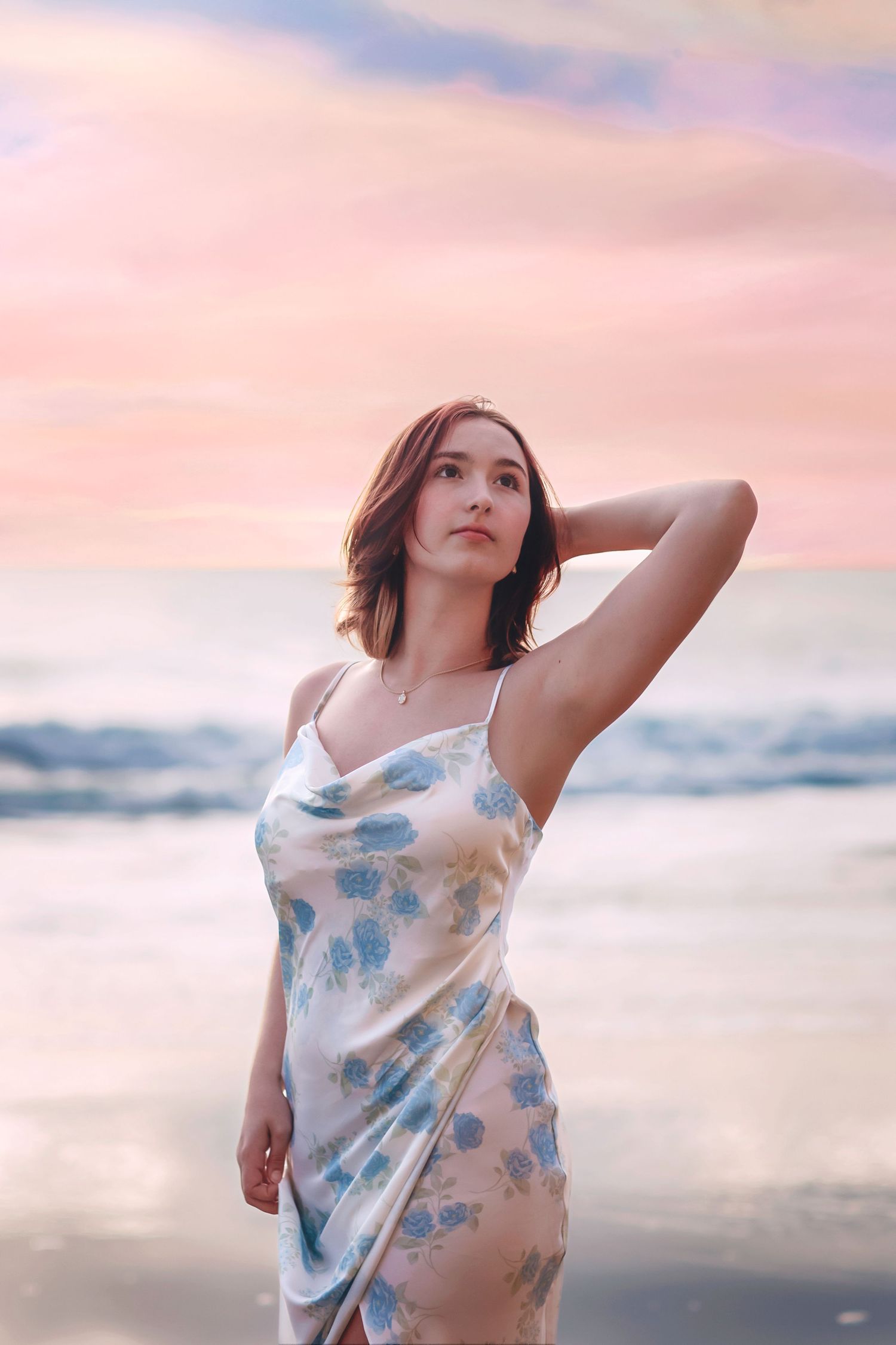 Young woman in floral slip dress poses gracefully on beach against soft pink sunset sky.