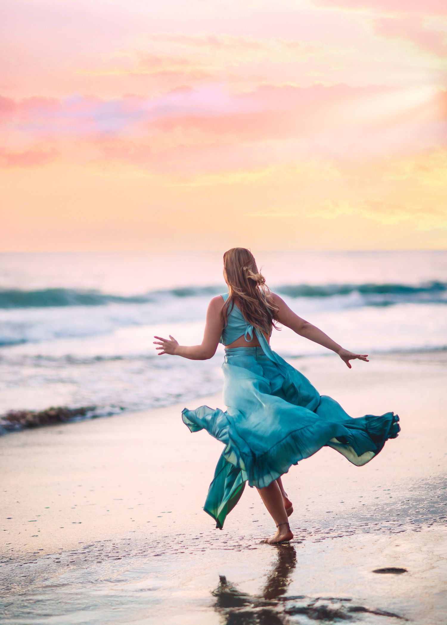 Woman in teal dress dancing on beach at colorful sunset.