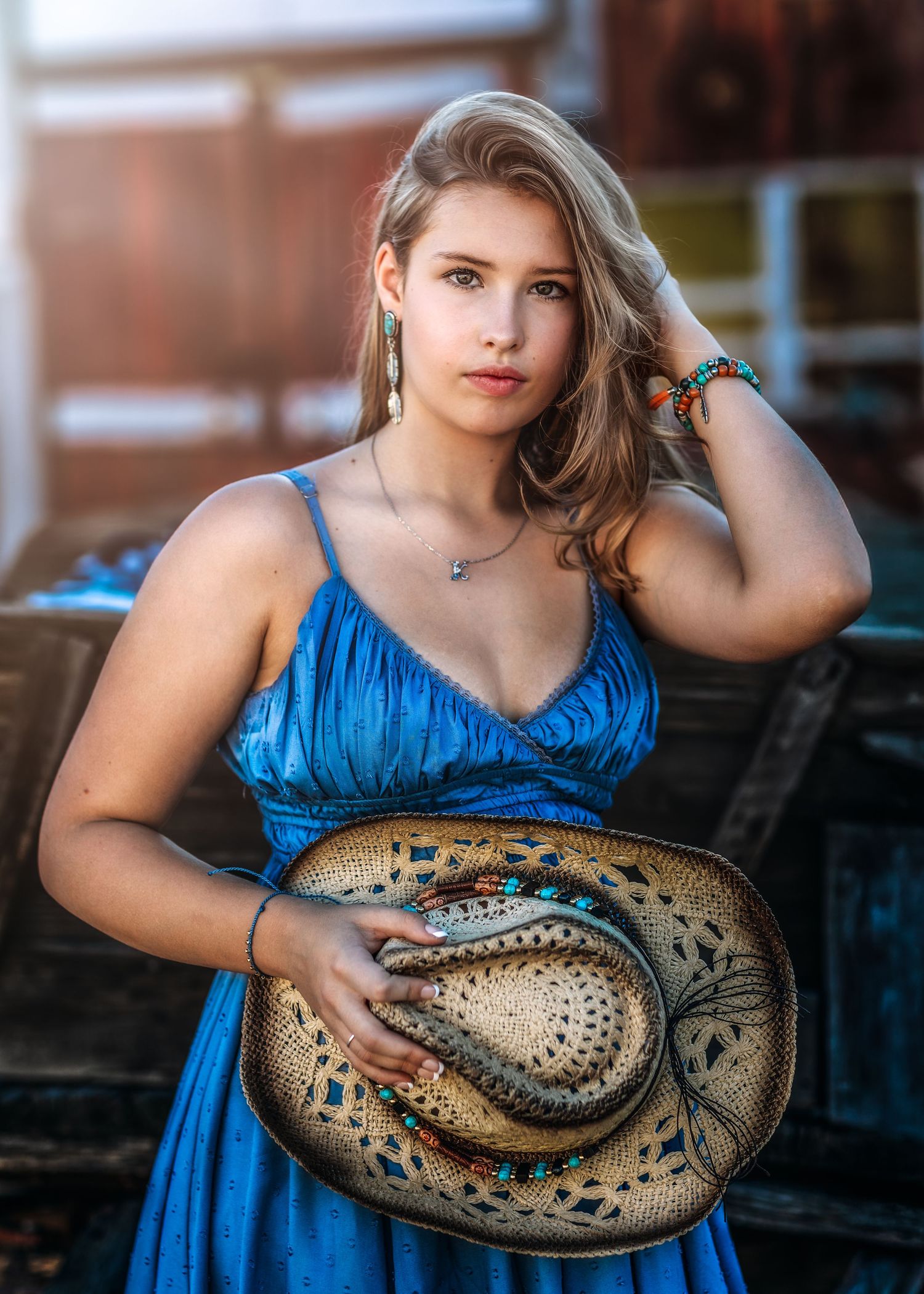 Young blonde woman in blue dress holding decorated cowboy hat with colorful beaded jewelry outdoors.