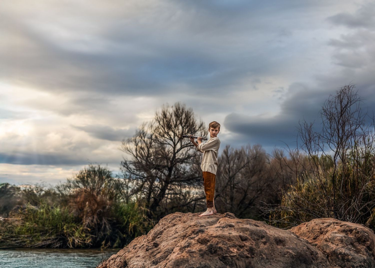 Child standing on large rock outdoors gesturing toward scenic landscape under cloudy sky.