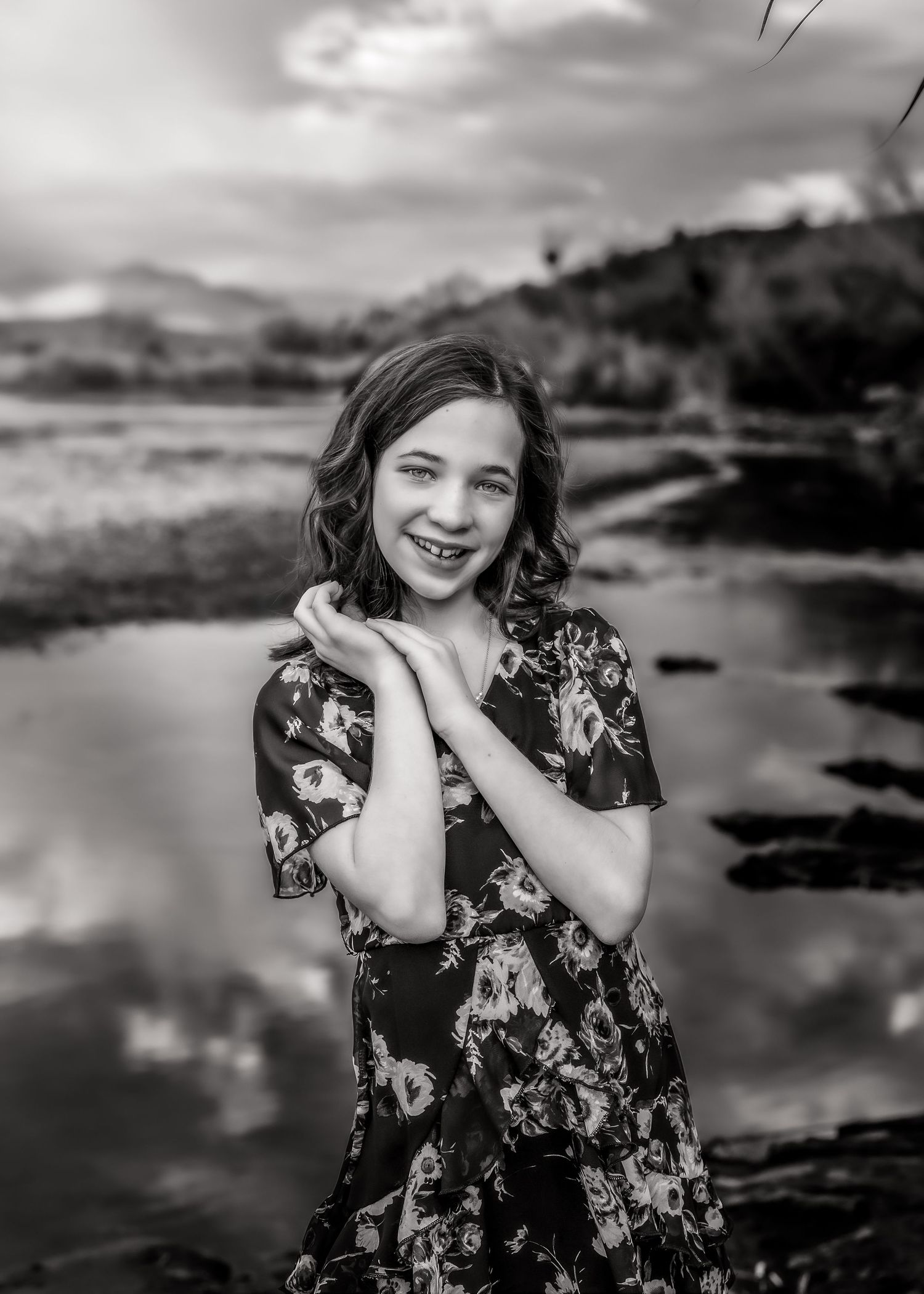 Black and white portrait of a smiling girl in a floral dress standing near a reflective pond.
