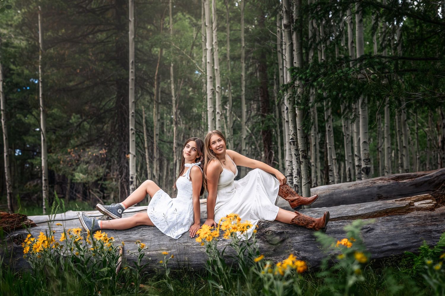 Two women in white dresses sit on a log in a lush forest surrounded by yellow wildflowers.