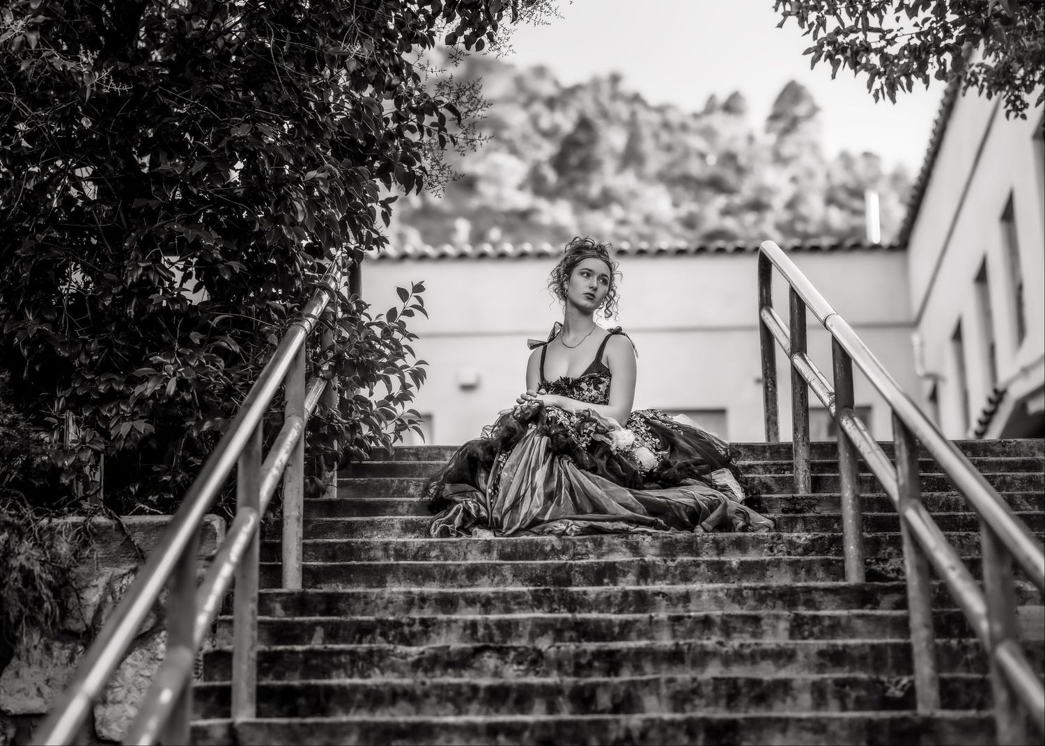Woman in elegant gown sitting on outdoor stone staircase surrounded by lush trees, black and white photography.