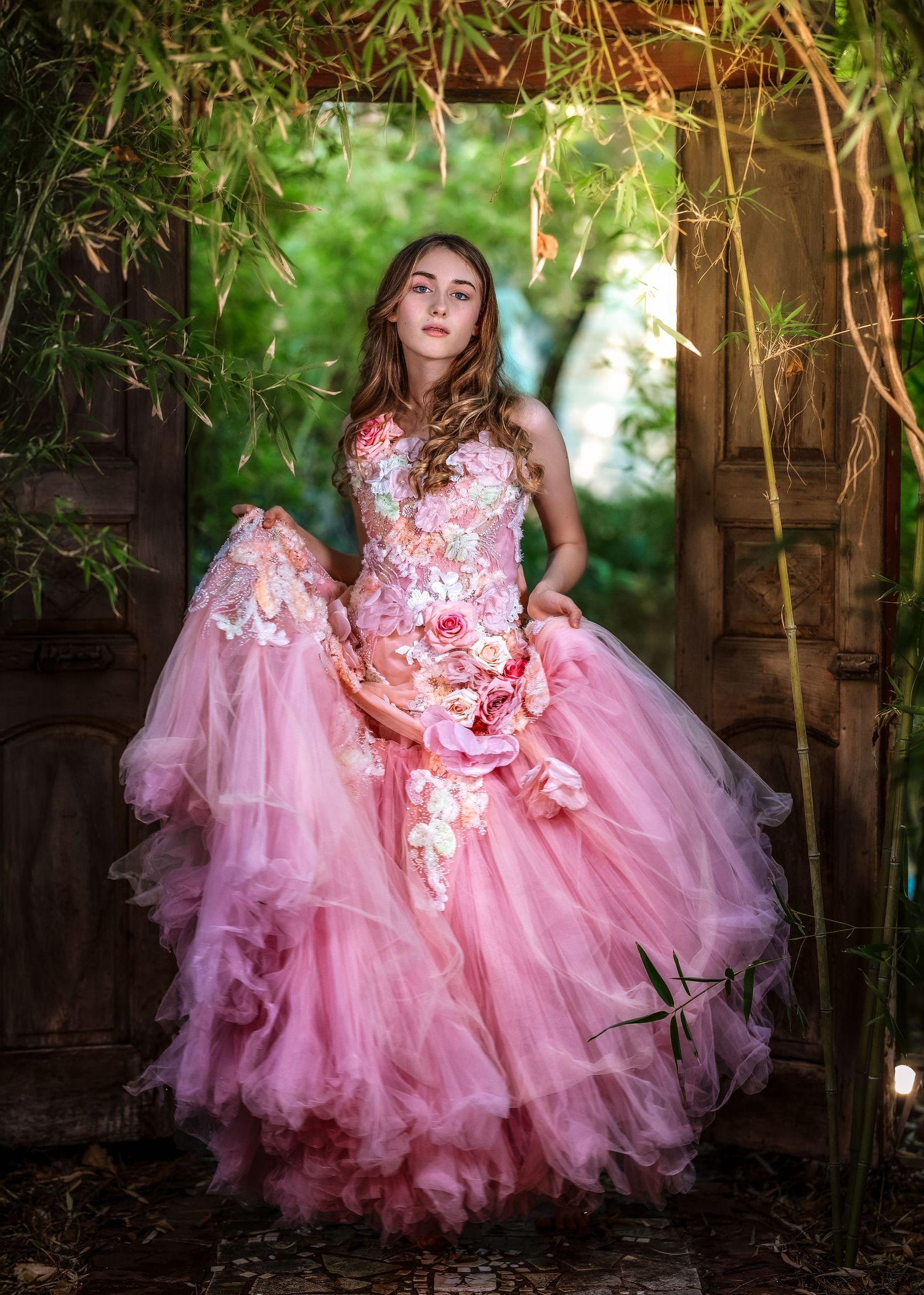 Girl in pink floral applique tulle gown stands in a bamboo archway doorway in a magical garden.