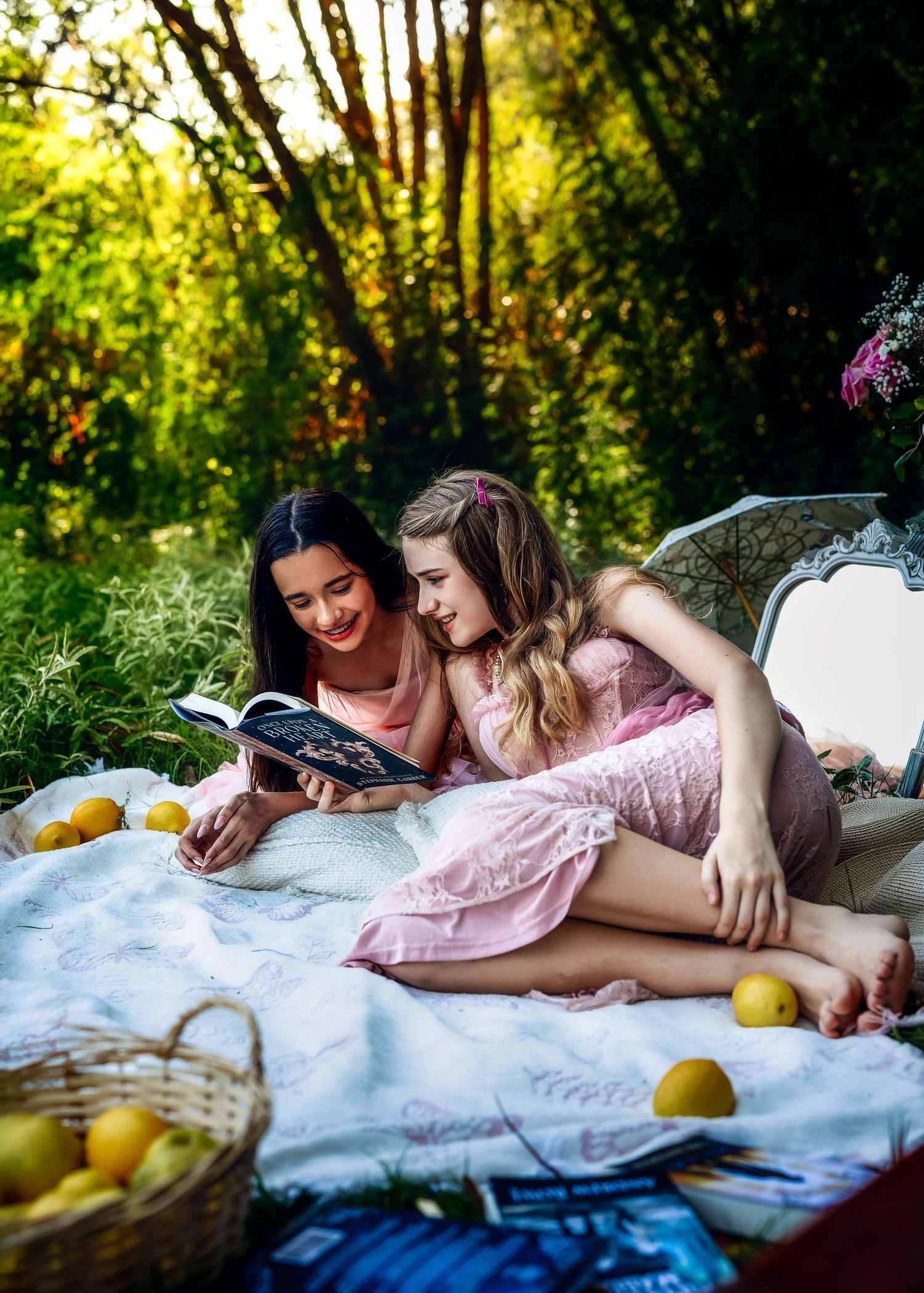 Two girls in pink dresses lie on a blanket outdoors reading a book surrounded by lemons and greenery.