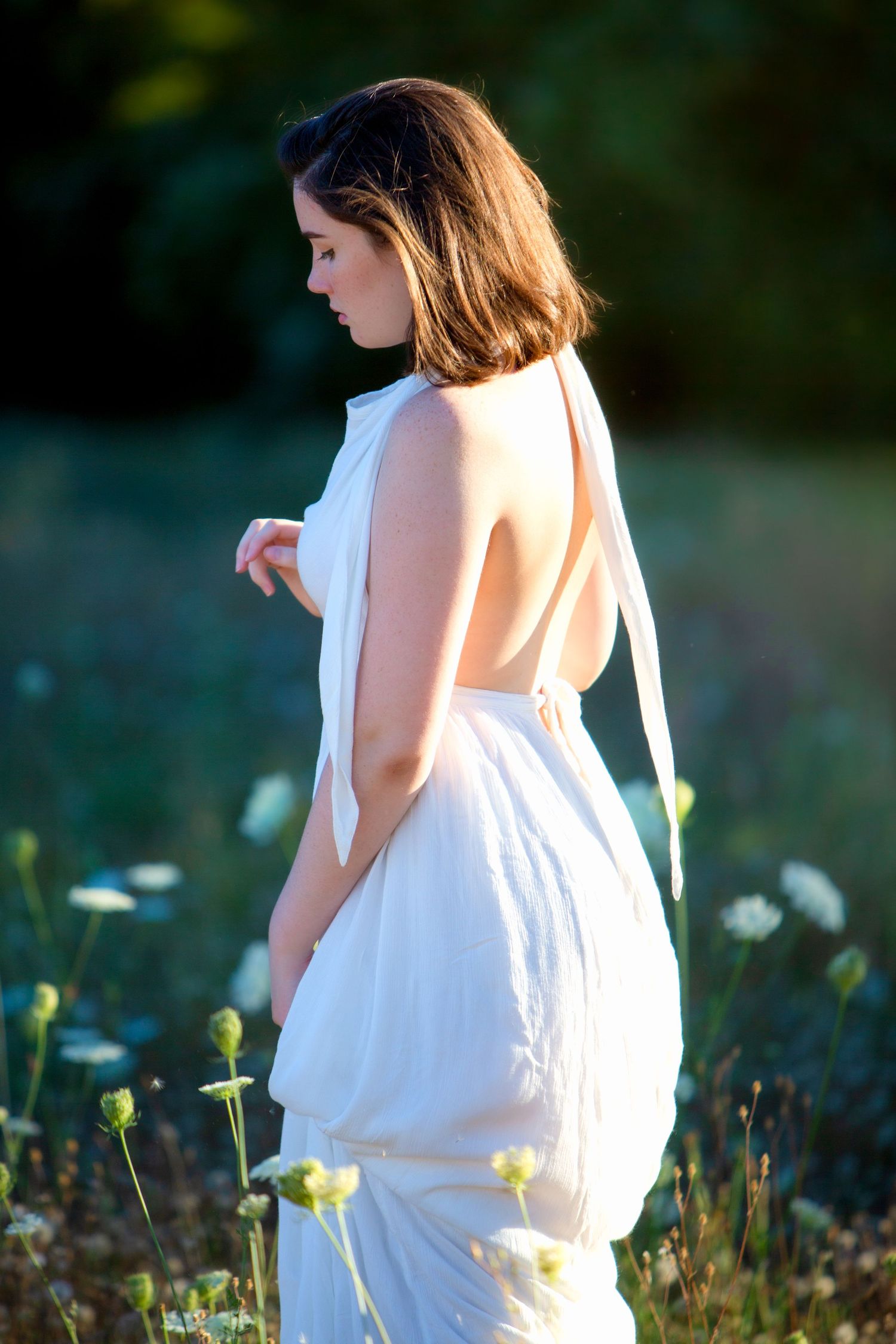 Young woman in white backless halter dress standing among wildflowers in soft evening light.