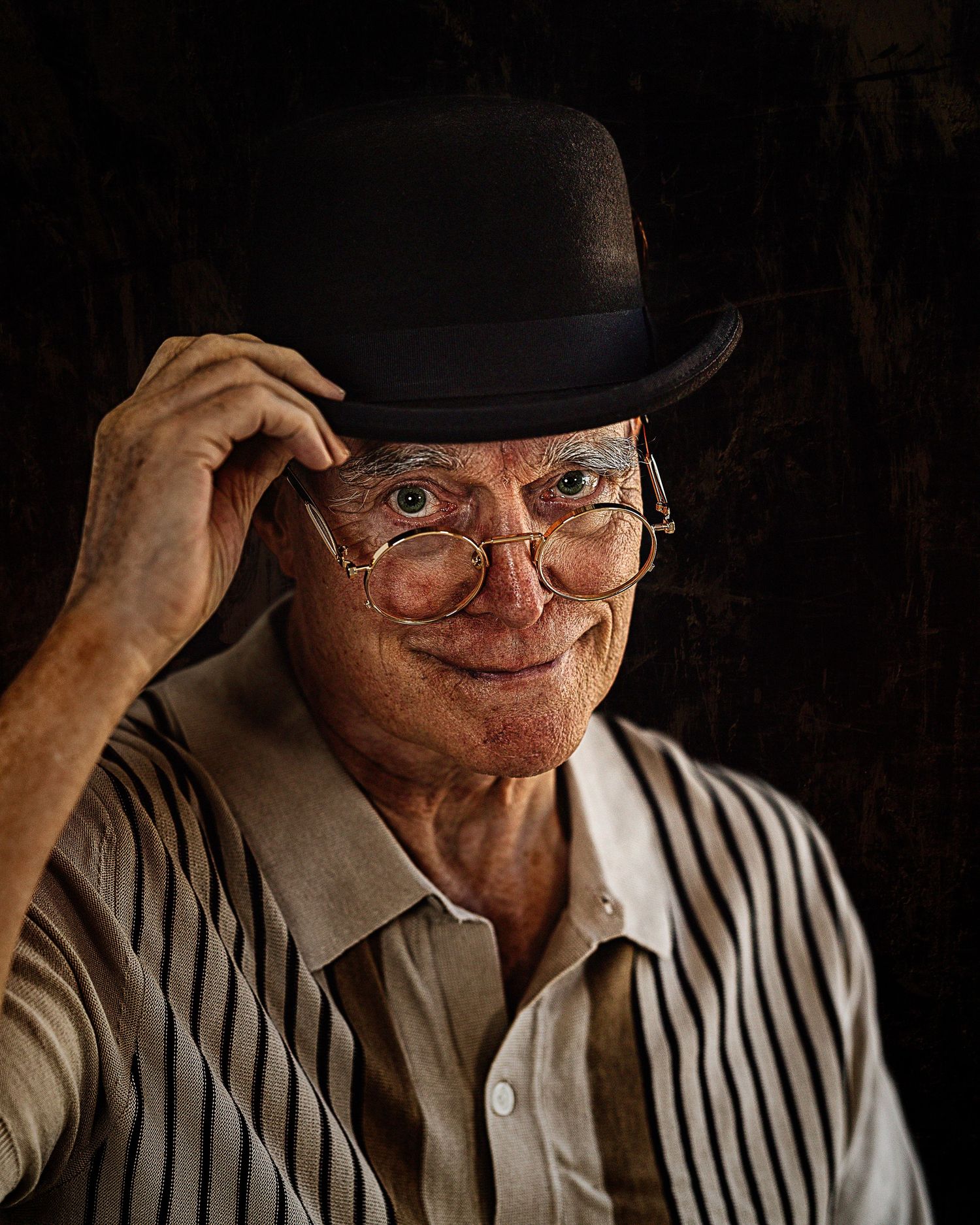 Elderly man smiling while tipping his black bowler hat, wearing round glasses and striped shirt.