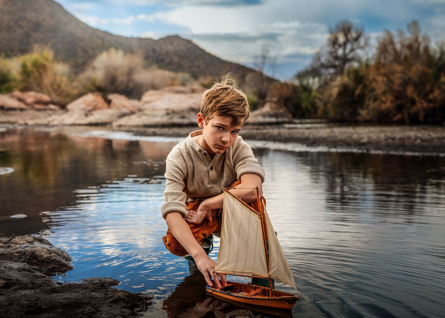 Boy crouching by calm water carefully placing a wooden sailboat into the river.