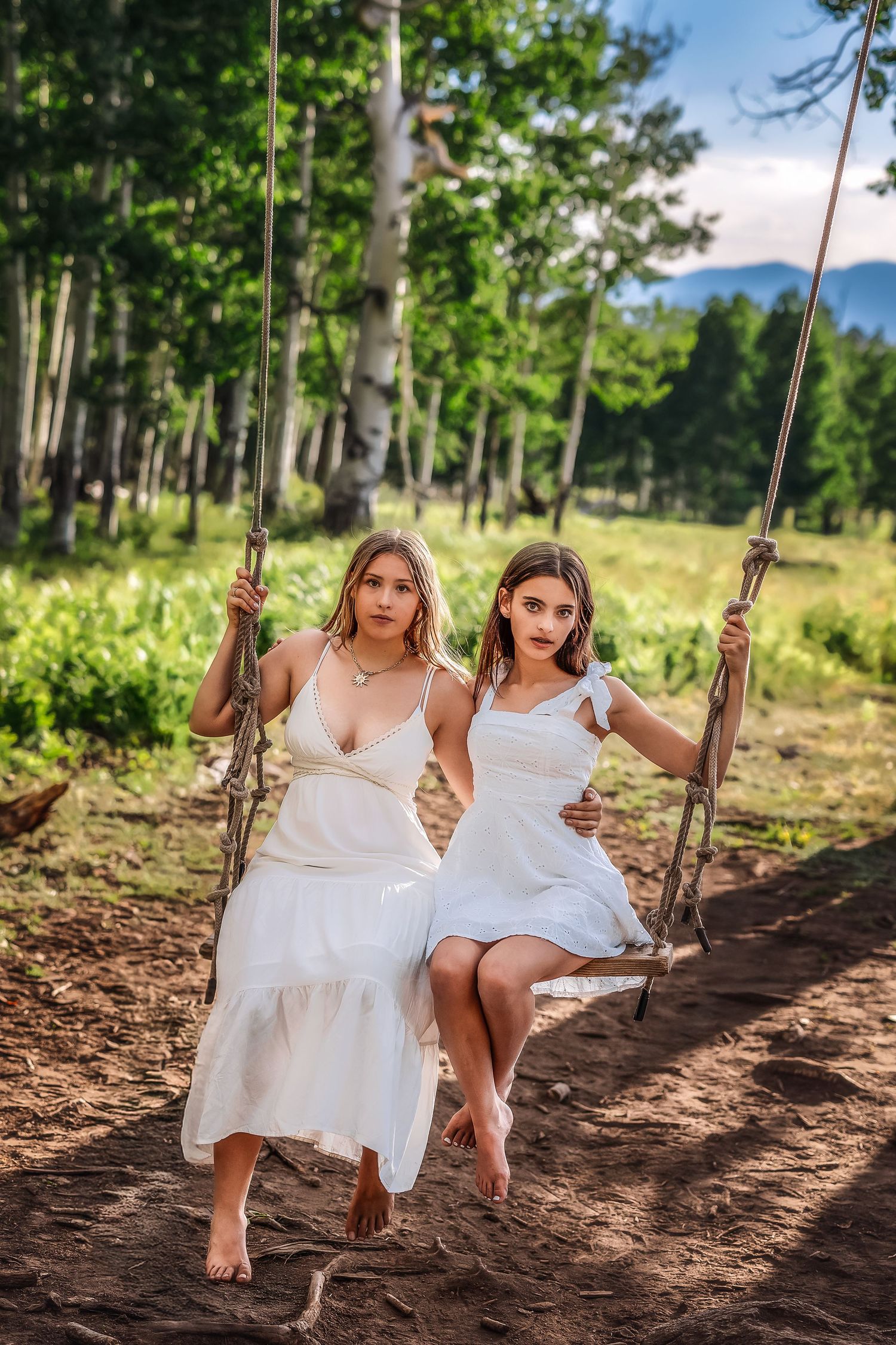 Two young women in white dresses sitting on rope swings in a lush green forest setting.