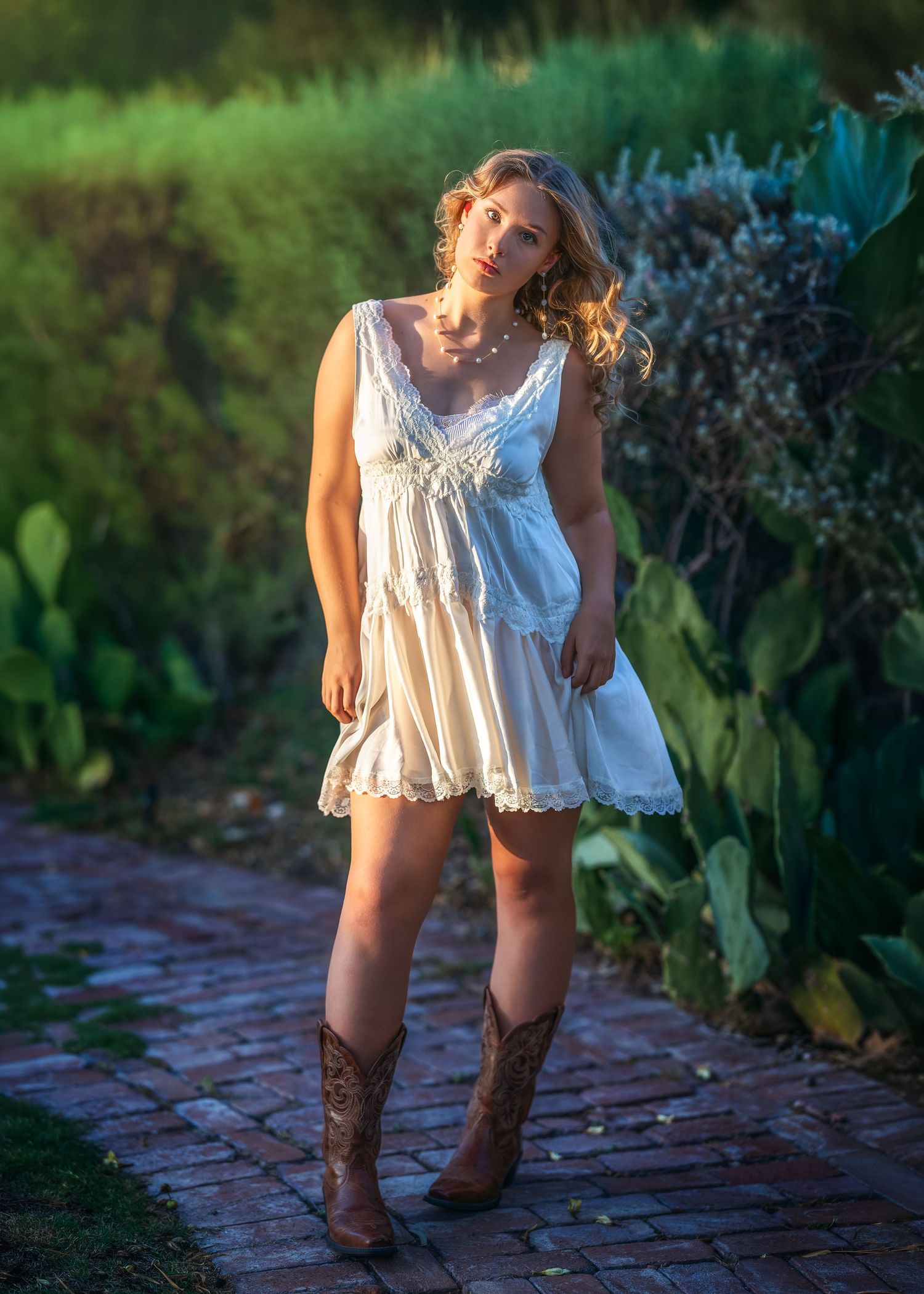 Young woman in white lace mini dress and brown cowboy boots standing on brick path surrounded by lush greenery.