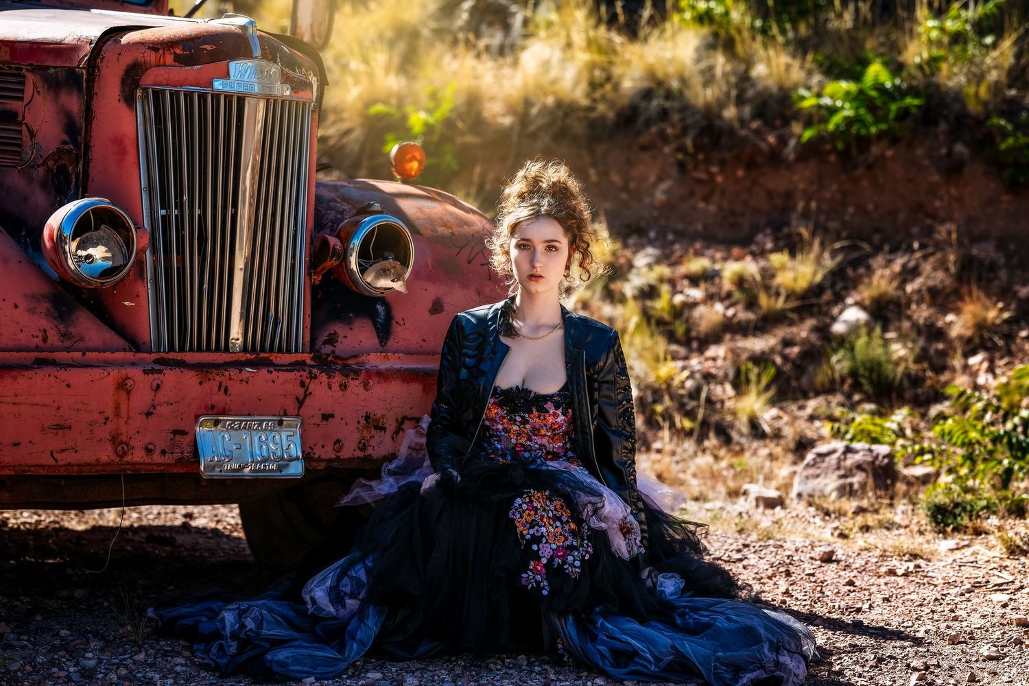 Woman in black floral gown sitting beside vintage red truck outdoors.