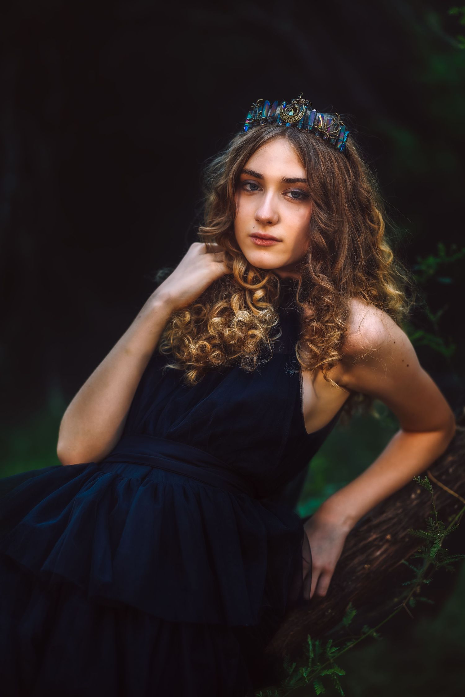 Young woman in black dress and decorative crown poses gracefully outdoors in a dark forest setting.